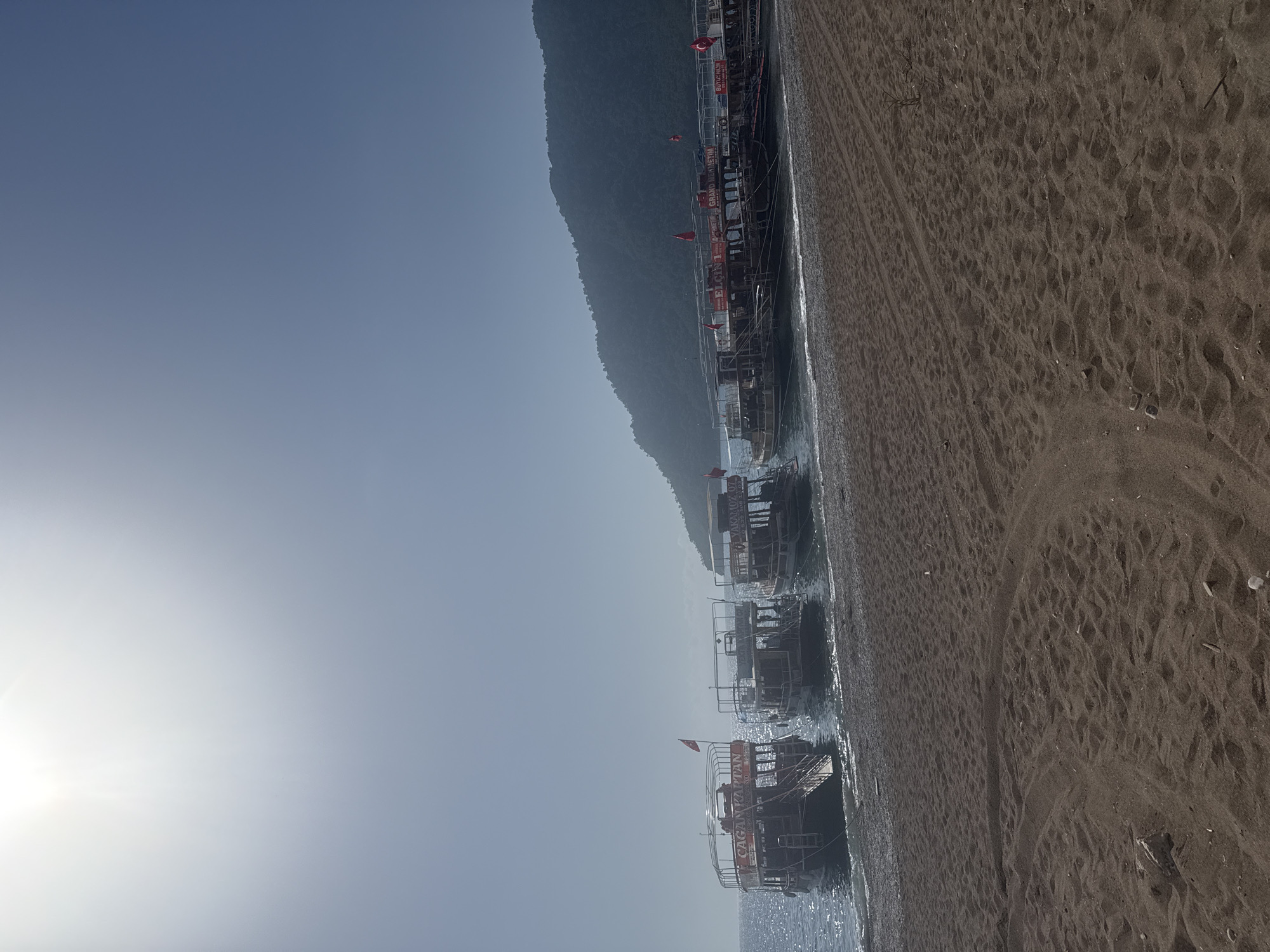 The image captures a coastal scene in Kumluca, Turkey. Several tour boats are moored along the shoreline. The boats are decorated with Turkish flags. A mountain rises in the background. The beach is dark and sandy. The sky is clear and the sun is bright.