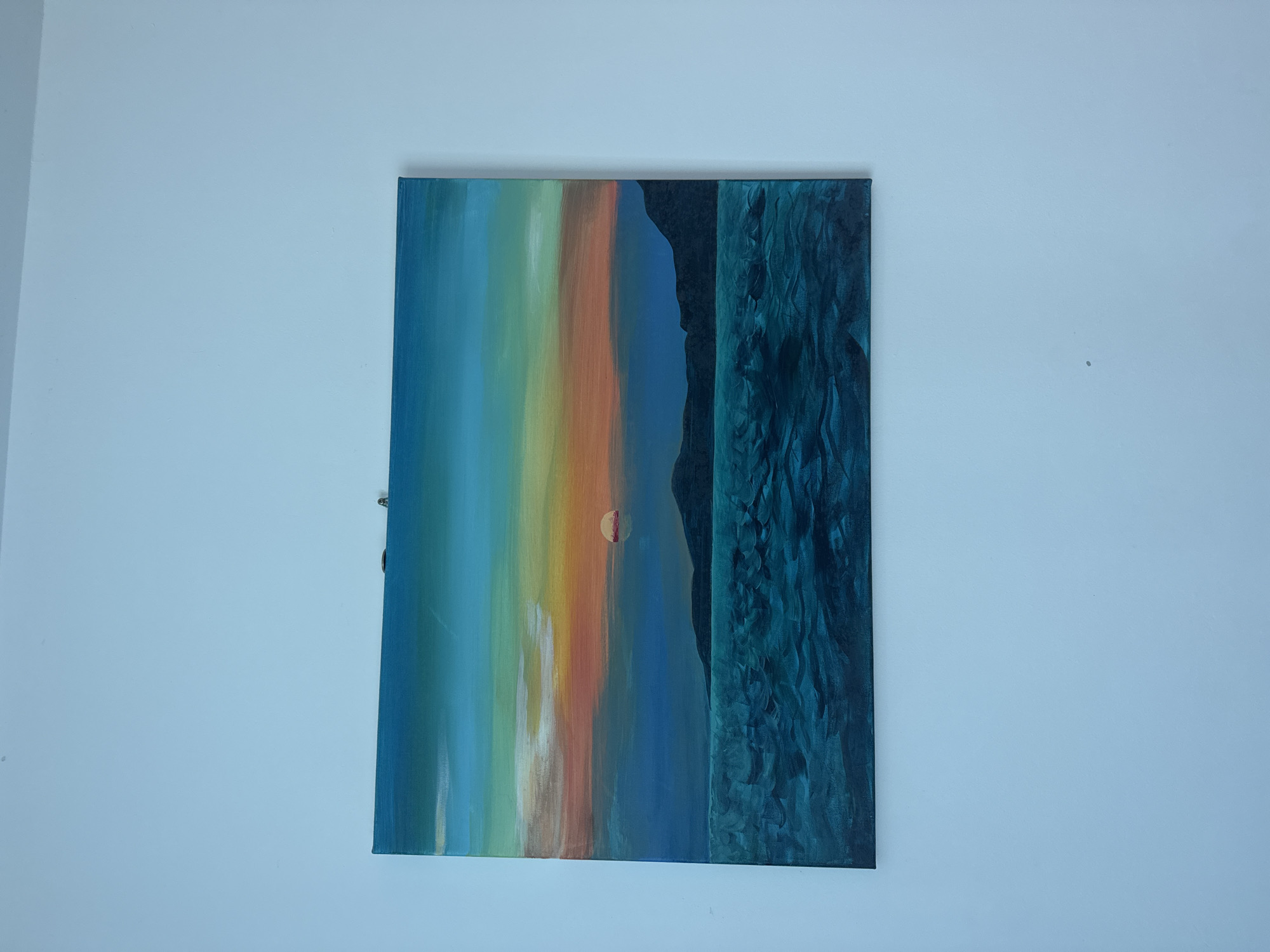 A painting hangs on a light blue-grey wall. The painting is composed of vertical stripes of varying colors, ranging from blue and green to orange and brown. There is a dark blue, textured area to the right of the colored stripes, separated by a solid blue-grey vertical section. A small, orange circular form is visible in the center. The painting appears to be mounted with a hook system.
There is no evidence to indicate that this image was captured in Pattaya City, Thailand.