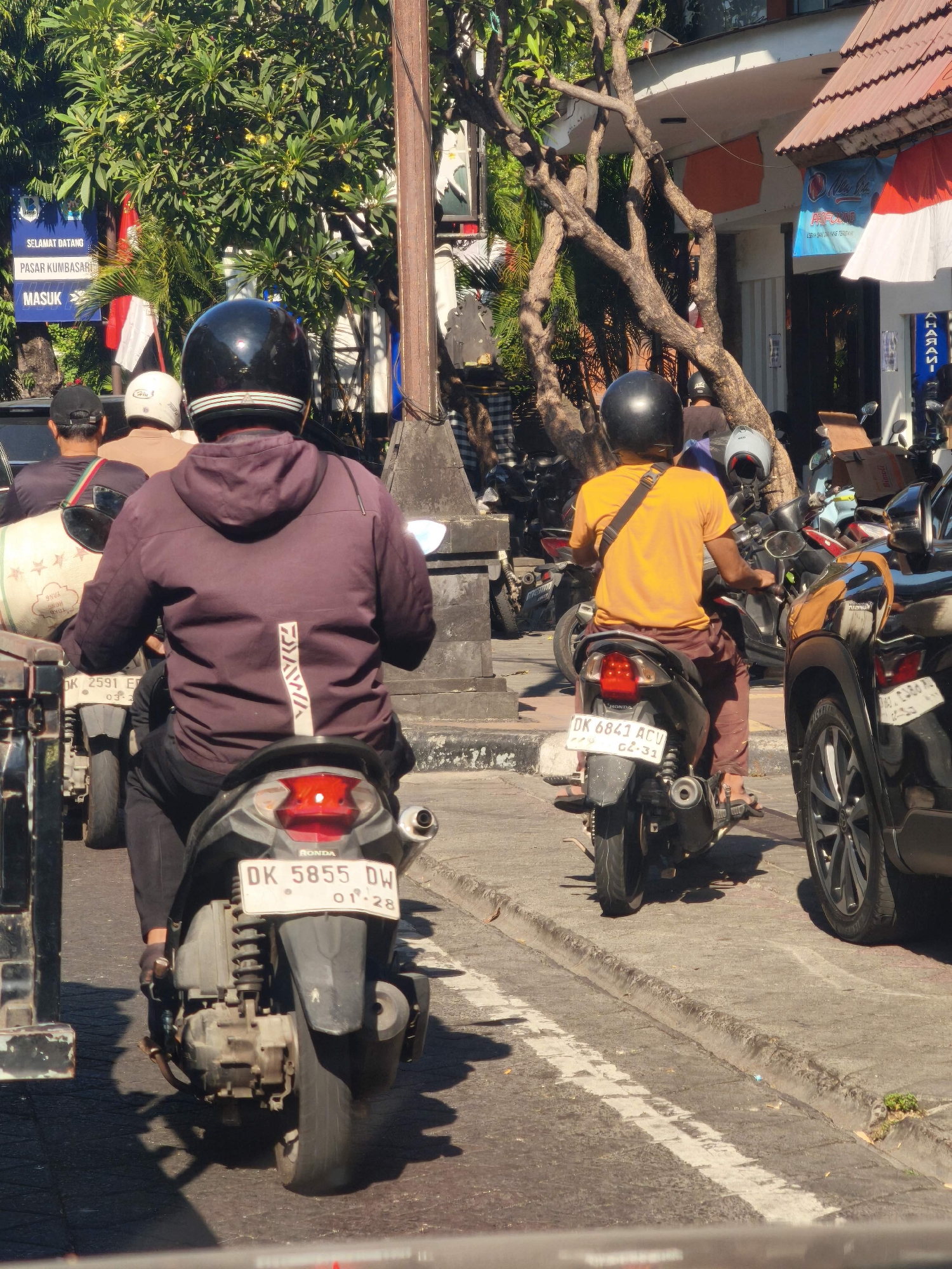 A medium shot captures a street scene in Denpasar, Indonesia, on a sunny day. The foreground is dominated by the rear view of two motorcycles and their riders. The motorcycle on the left is dark gray with a red taillight and a white license plate that reads "DK 5855 DW 01-28". The rider wears a dark purple hooded jacket and a black helmet. Beside this motorcycle, another dark gray vehicle, possibly a car, is partially visible on the right, showing its rear wheel and part of its body.
The motorcycle on the right is black with a white license plate reading "DK 6843 ACV 02-31". The rider, wearing an orange t-shirt and brown pants, has a black helmet. Several other motorcycles are parked or moving in the background, along with a few people on foot or on scooters. The environment includes a sidewalk, street, trees, and buildings with signs. One sign visible on the left reads "SELAMAT DATANG PASAR KUMBASARI MASUK". The overall atmosphere suggests a bustling urban environment.