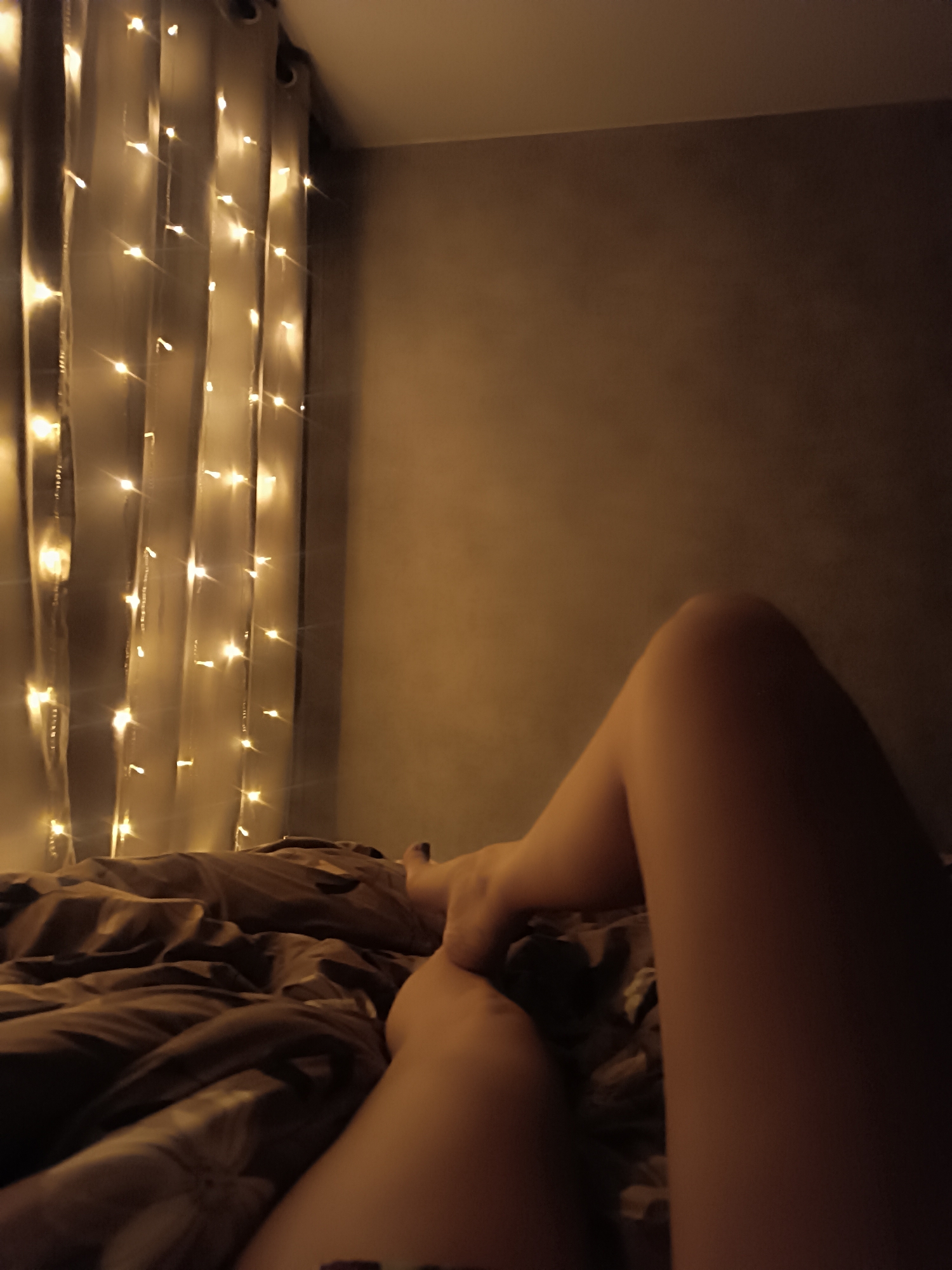 The image shows a view from a bed looking towards a curtain with lights. A person's legs are visible, with the feet near the middle and the legs extending towards the bottom right. The bed has brown bedding. Behind the curtain is a wall. The location is a bedroom.