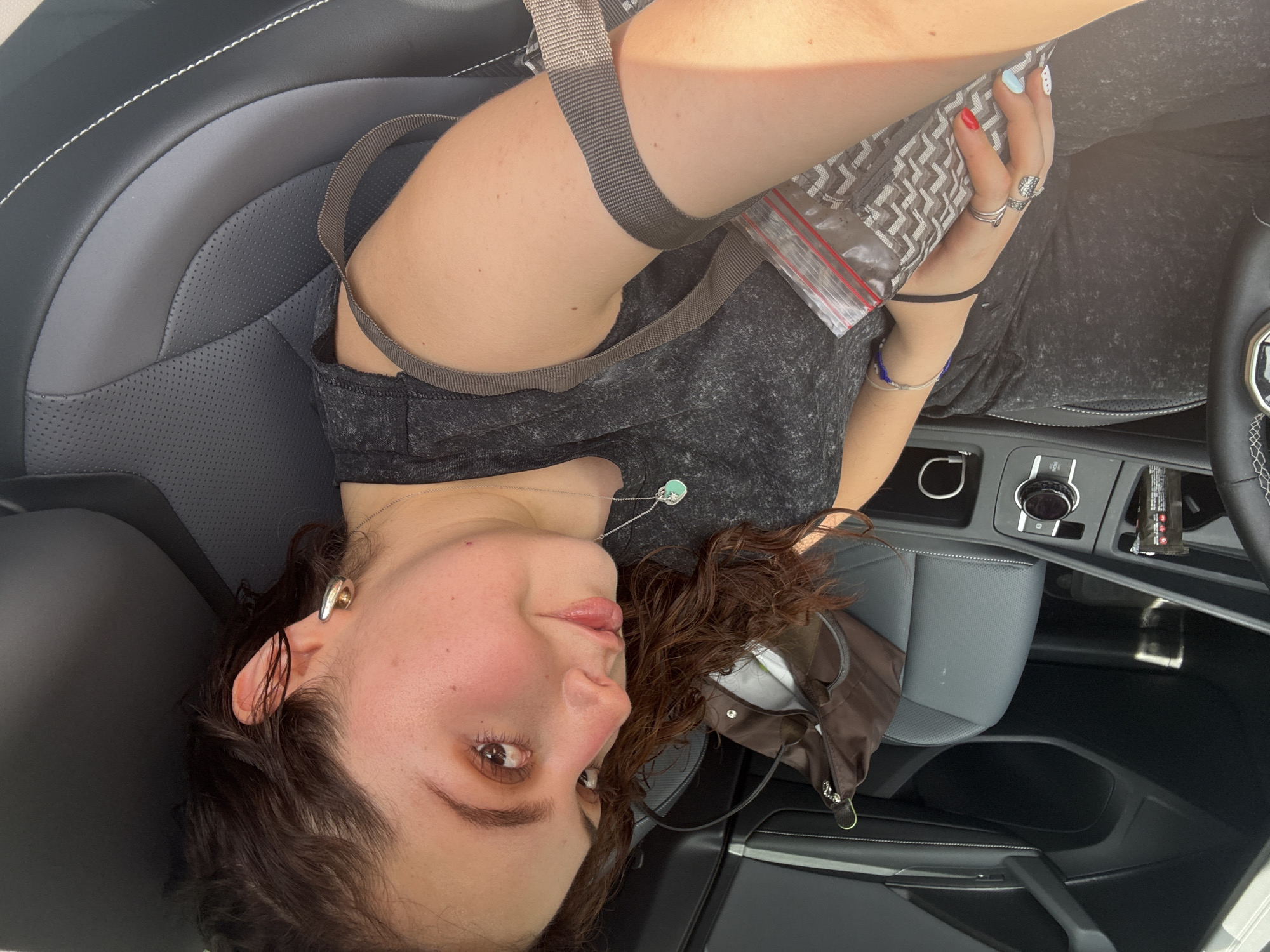 A sideways, high-angle close-up photograph captures a young woman reclining in the passenger seat of a modern car. She has medium-dark, curly hair, and her head is positioned on the left side of the frame, looking towards the camera with a calm, slightly pensive expression, featuring soft, rosy blush on her cheeks and visible eye makeup.
She is wearing a dark gray or black marled t-shirt or tank top. A thin silver chain with a light blue-green pendant hangs around her neck, and a silver ear cuff adorns her right ear. Her left hand, adorned with multiple silver rings, holds a clear plastic bag with a red zipper closure, containing a gray and white patterned item, possibly fabric. Her fingernails are painted in various colors and designs, including red, light blue, and white with dots. A black elastic hair tie is on her left wrist.
The car's interior features black and gray leather seats with white stitching, indicating a contemporary vehicle. A brown strap, likely a car seatbelt, crosses her upper body. Part of the car's dashboard, including control knobs and a section of the steering wheel, is visible on the right. A small brown bag or pouch rests on the car seat near her left side. Bright light, possibly sunlight, illuminates her right arm and hair, suggesting daytime. The general atmosphere is relaxed. No visible text can be clearly read in the image. The location context provided for the image is Mexico City, Mexico.
