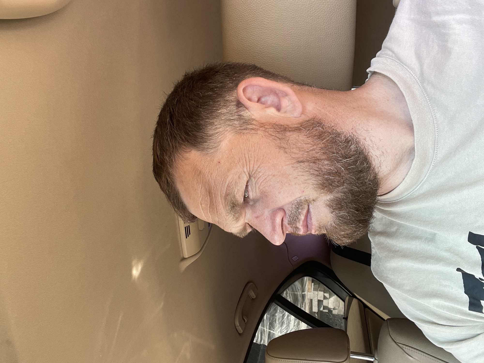 A man is inside a car. The interior of the car is beige. The man has short brown hair, a beard, and is wearing a light-colored t-shirt. The scene appears to be inside a vehicle due to the proximity of interior car elements such as the headrest, side panel, and window. There is no indication of the location being in Krasnodar, Russia.