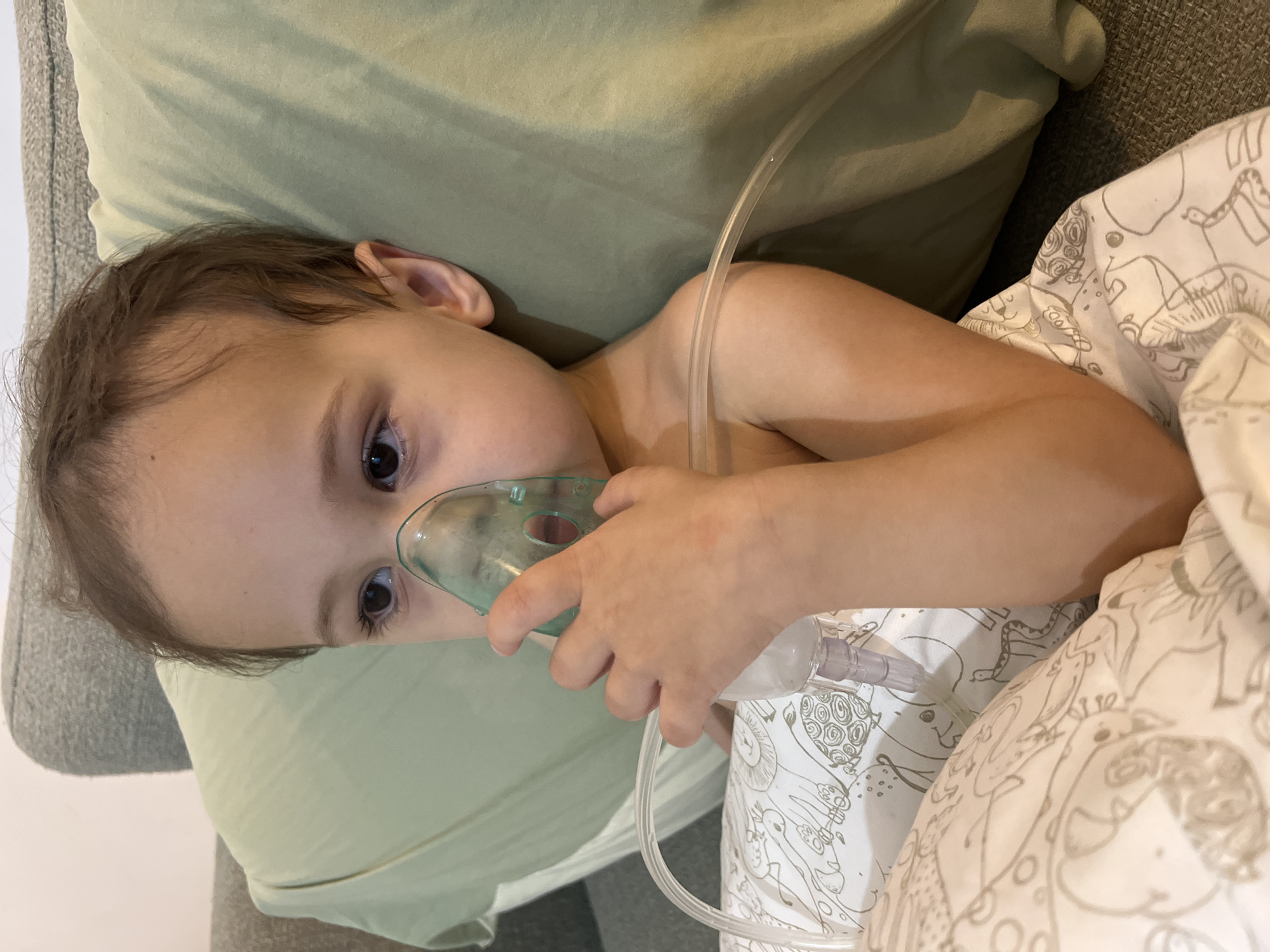 A young child, approximately a toddler, is depicted in an indoor setting within Kiryat Bialik, Israel. The child, shirtless with light brown hair and dark eyes, is holding a green nebulizer mask over their mouth and nose with their right hand. A clear plastic tube connects the mask to a small, clear medication cup, positioned over a white blanket with light grey cartoon animal and abstract patterns. The child is reclining against a light green pillow, which rests on a grey, textured sofa. A pale, off-white wall is visible in the upper left background. The child's gaze is directed towards the right side of the frame.