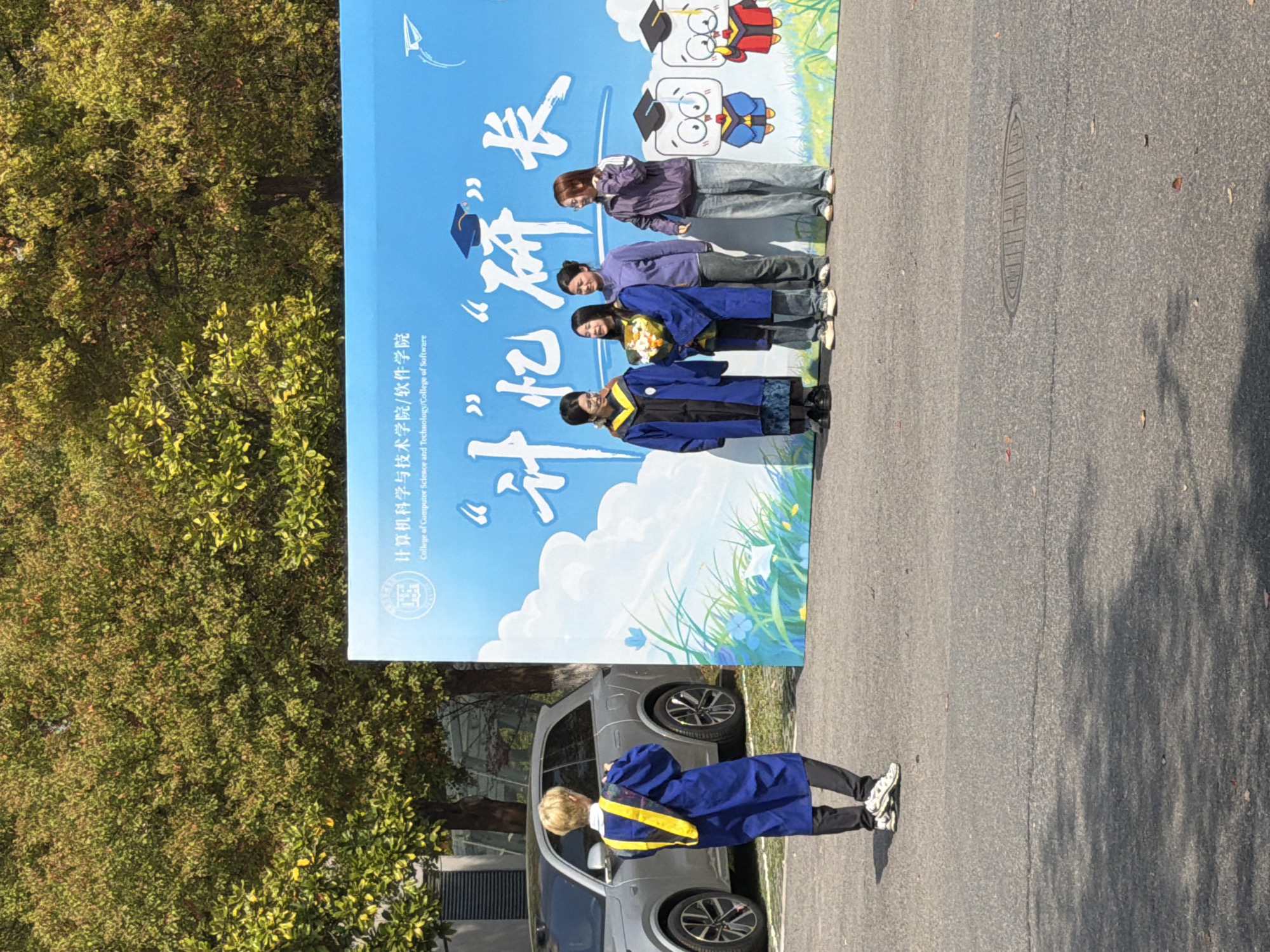 Here is a description of the image:
The image shows an outdoor scene, likely a campus area, based on the presence of trees and pavement. The photograph features a vertical banner with a blue background and white calligraphy and images of figures in graduation caps. Four people are positioned near the banner, appearing to have taken a photograph with it. In the foreground, a person in graduation attire (a blue gown with a yellow sash) stands near a parked silver car, likely posing for a picture as well. The location might be in China based on the text on the banner and the style of dress. The city Jiangning District, China could possibly be the city because the title asked to include it in the description of the image.