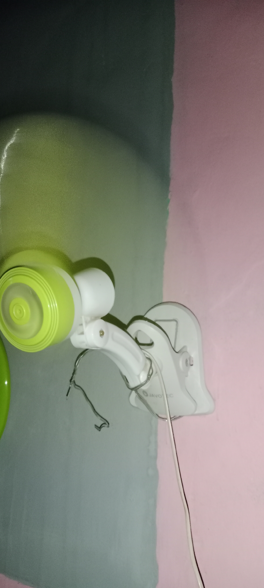 The image shows a close-up, angled shot of a white baby monitor with a green dome-shaped camera unit attached to a flexible white arm. The monitor is clipped onto a wall that is painted in two distinct colors: a darker gray-green on the left and a lighter pink on the right, with a clear, vertical dividing line between them. The monitor itself has the brand name "JAVO" visible on its base. Wires are attached to the monitor, one of which appears to be metallic and coiled. The lighting suggests an indoor setting, possibly a nursery, with a soft glow illuminating the green camera unit. The image does not contain any information about the city of Subang, Indonesia.