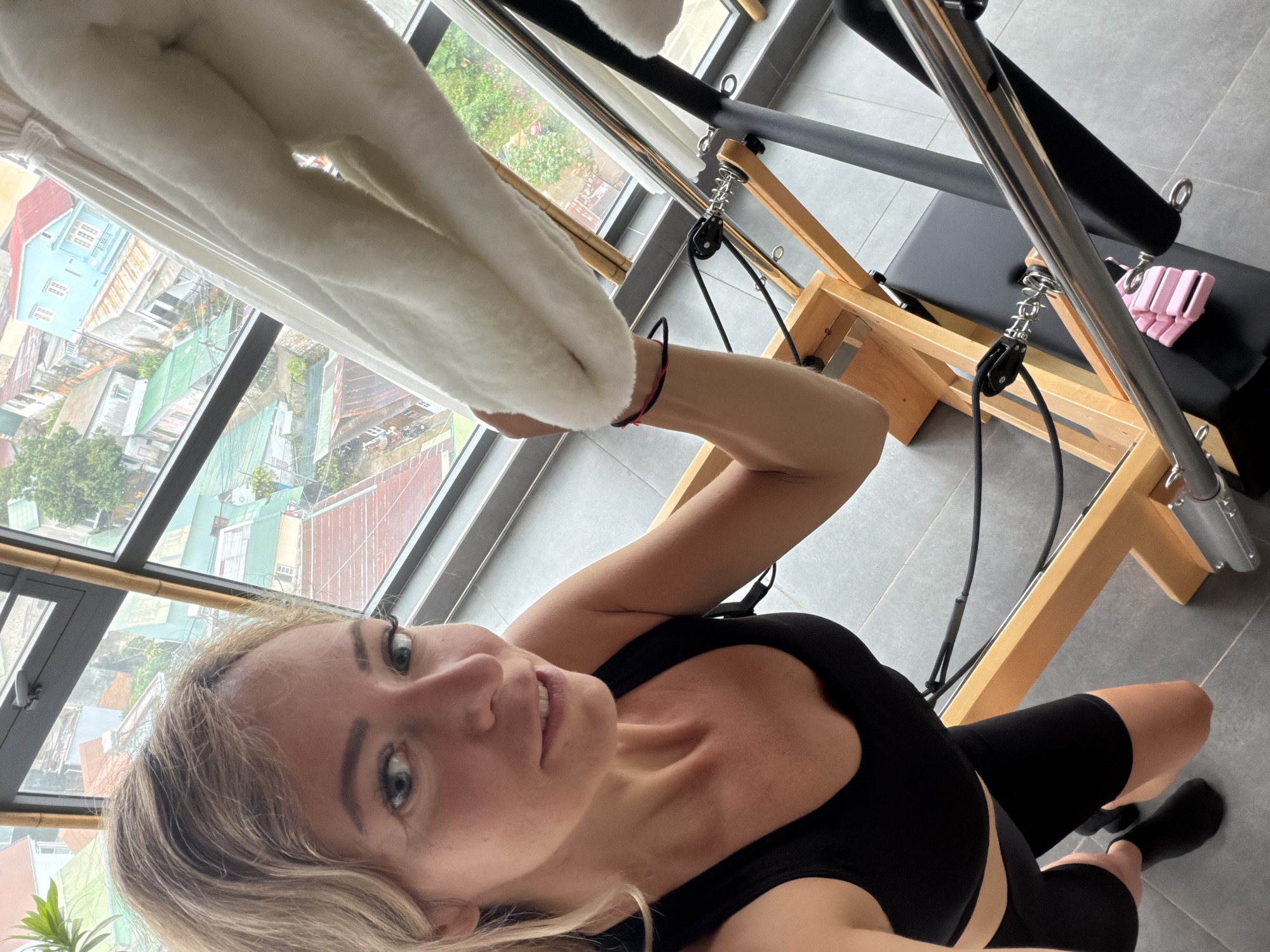 A woman with blonde hair, wearing a black sports bra and black shorts, is in a room with a large window overlooking a city. The room appears to be a Pilates studio, as a Pilates reformer machine is visible. The woman is looking at the camera and smiling. The view from the window shows a dense urban landscape with red-tiled roofs and some green trees. The architecture suggests a Southeast Asian setting, possibly Da Lat, Vietnam, given the style of the buildings and the lush vegetation. The overall impression is of a bright, modern space combined with a vibrant urban environment.