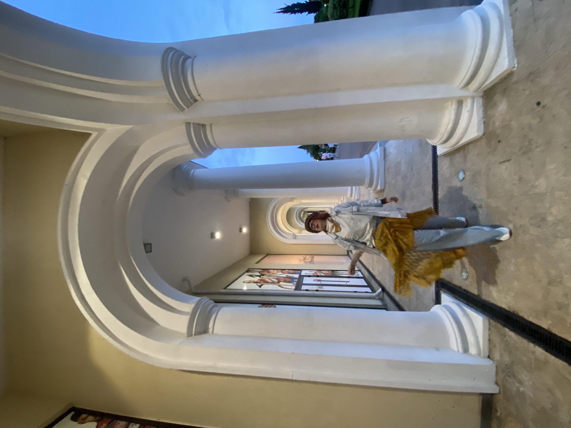 The image depicts a person standing in a colonnade or arched walkway. The walkway is characterized by its classical architecture, featuring white columns and arches. Artworks are visible along the walkway's walls. The person is wearing casual clothing and appears to be in motion. The location is likely Sochi, Russia.