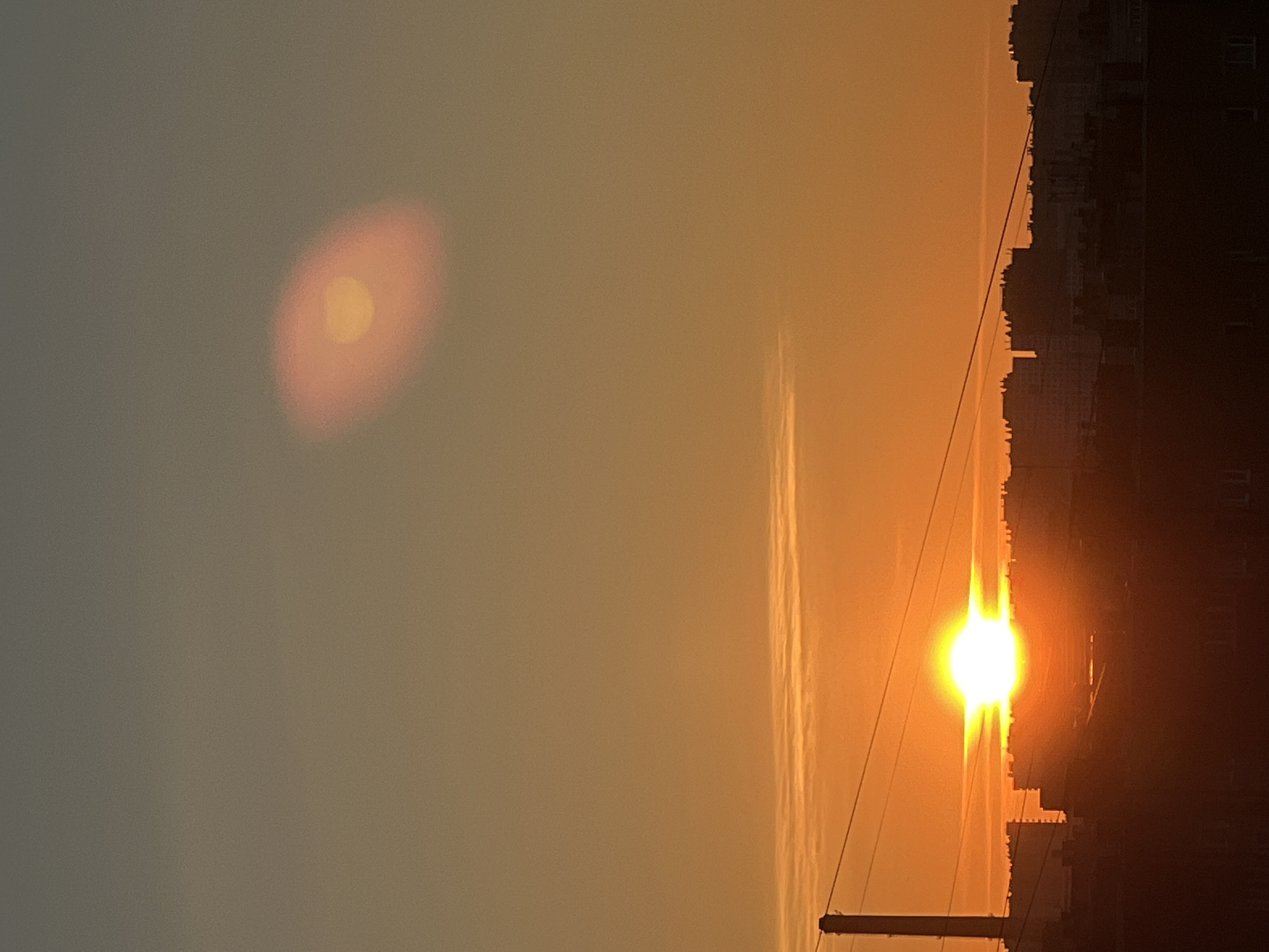 This is a vertical, full shot photograph of a sunset over the city of Sinyavino, Russia. The sky is a gradient of warm orange and yellow hues, with a hazy, slightly pinkish oval shape visible in the upper right quadrant. A bright, intense sun is low on the horizon, positioned in the center of the frame, emitting a strong glare. Silhouetted buildings form a jagged skyline across the lower half of the image, with a tall, narrow industrial chimney or tower visible on the left side. Thin power lines traverse the mid-section of the frame horizontally. The foreground consists of a dark, silhouetted roofline of buildings. There are no visible people or animals in the scene. The overall impression is of an urban sunset.