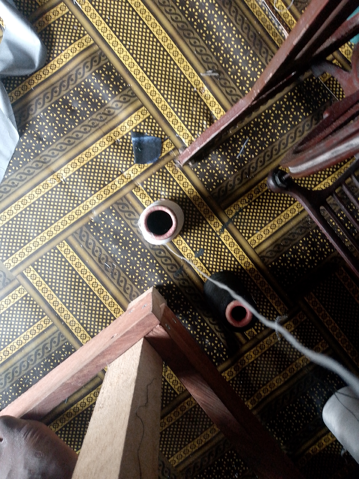 The image depicts an indoor scene with a patterned floor and wooden elements. A dark, patterned fabric covers a portion of the floor, while another section has a yellow and black geometric pattern. There is a small, dark square object on the floor near the patterned fabric. A white ceramic pot with a dark opening is present, along with a black cylindrical object with a red opening. Wooden framing, possibly from furniture or construction, is visible in the foreground and extends into the background. A light-colored piece of fabric or material is draped over a surface on the left side of the image. The lighting appears dim. The location is identified as Jauro Yinu, Nigeria.