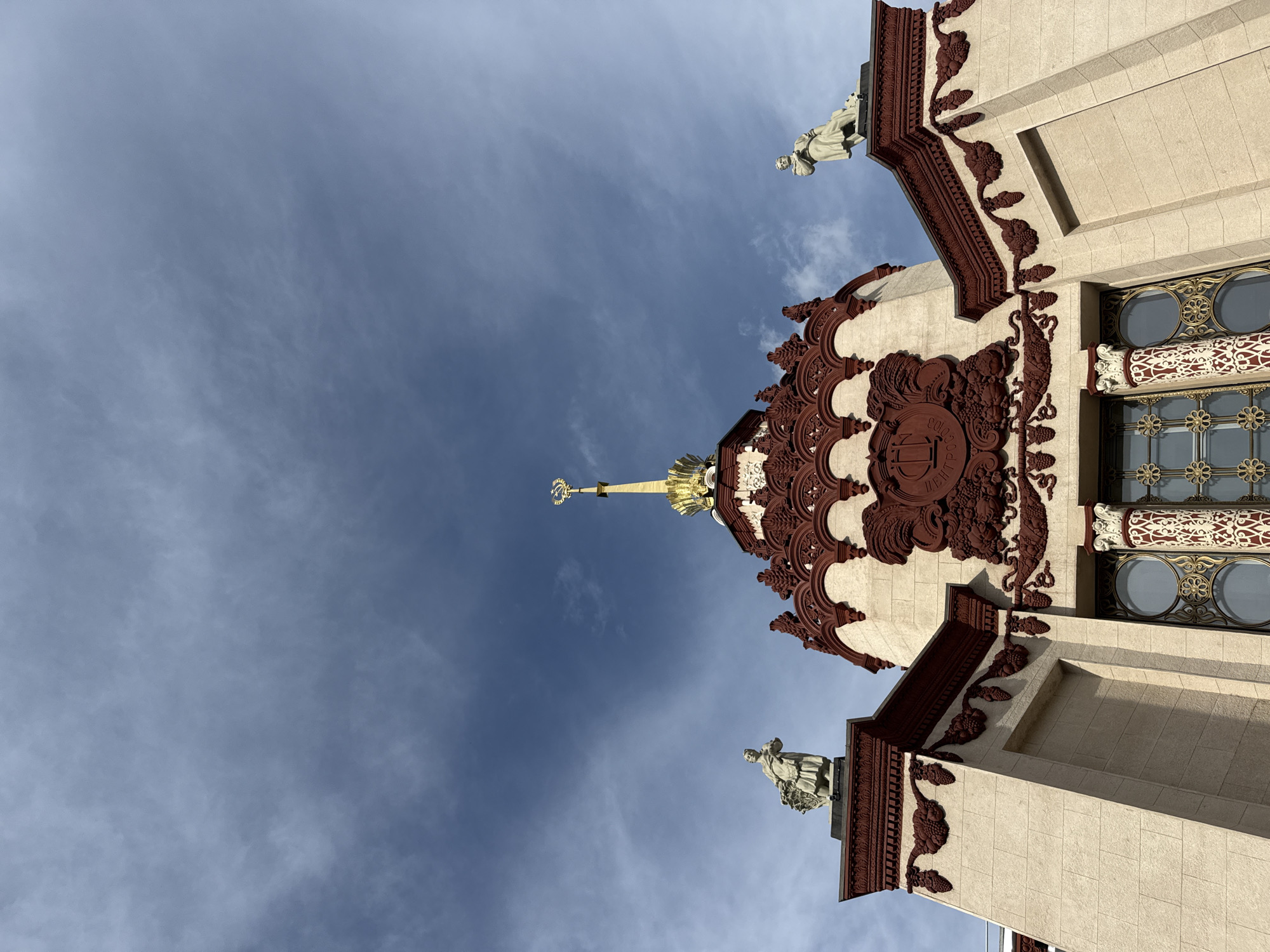 The image shows a low-angle view of the pavilion of the Central Union of Consumer Cooperatives (Tsentrosoyuz) at VDNKh (Exhibition of Achievements of National Economy) in Moscow, Russia. The building's ornate architecture features a light-colored facade with dark red decorative elements, including sculpted fruit and floral motifs, and arched windows with intricate metalwork. Two classical statues of figures holding baskets of produce stand on pedestals on either side of the upper portion of the building. The central spire is crowned with a golden hammer and sickle emblem, symbolizing the Soviet era. The sky above is a bright blue with wispy white clouds.