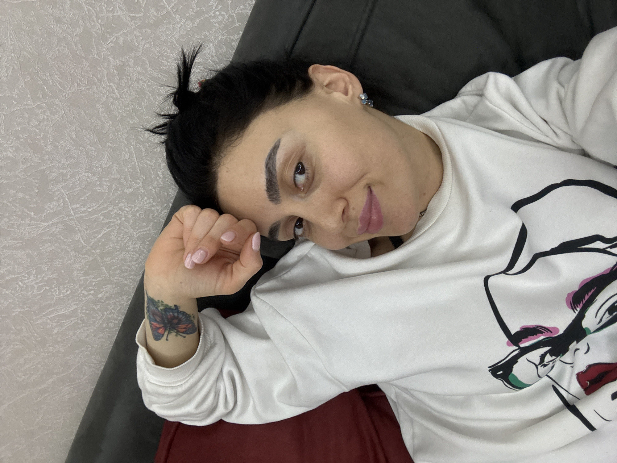A woman with dark hair pulled into a small bun at the top of her head, wearing a white sweatshirt with a drawing of a face, is lying on what appears to be a dark gray couch. Her left arm is bent and her hand is near her face. A butterfly tattoo is visible on her left wrist. The background is a light gray wall. No information is available to confirm whether this scene took place in Tbilisi, Georgia.