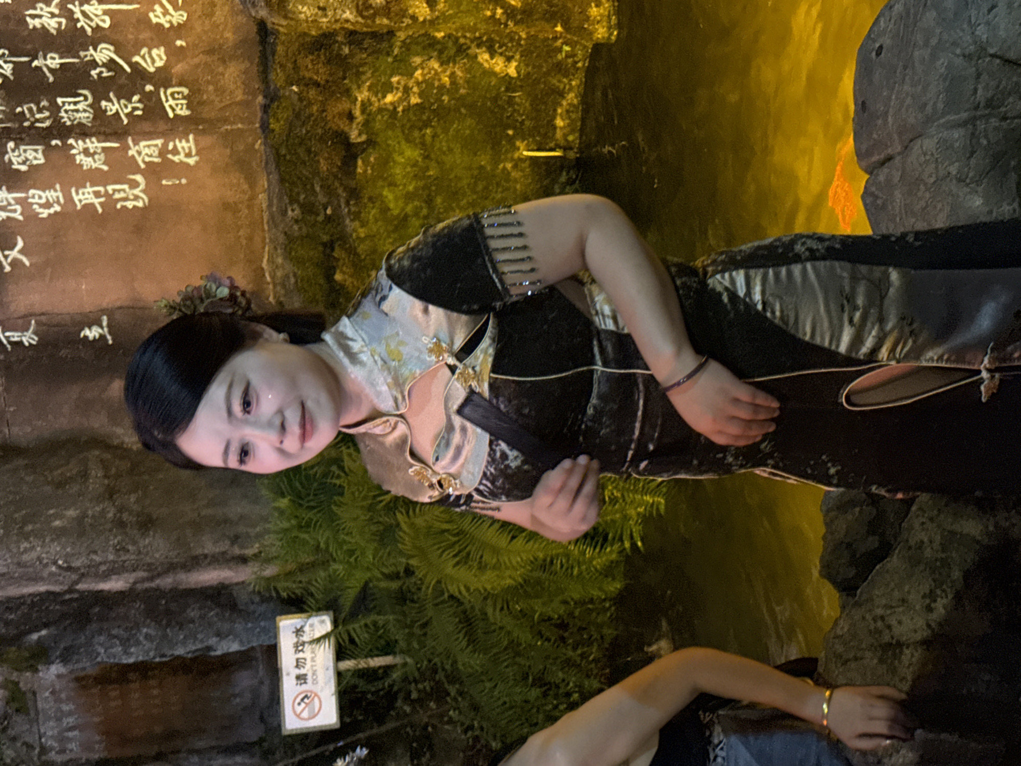 A woman is pictured in a traditional Chinese dress in Yuzhong District, China. The dress is black with silver accents and has a high collar. The woman has dark hair pulled back and is wearing makeup. She is standing in front of a rocky wall with Chinese calligraphy written on it. There is a sign on the wall that says "DON'T PLAY WITH WATER." In front of her is a body of water with yellow lighting. There is also foliage visible in the background. Another person is partially visible on the left side of the image.