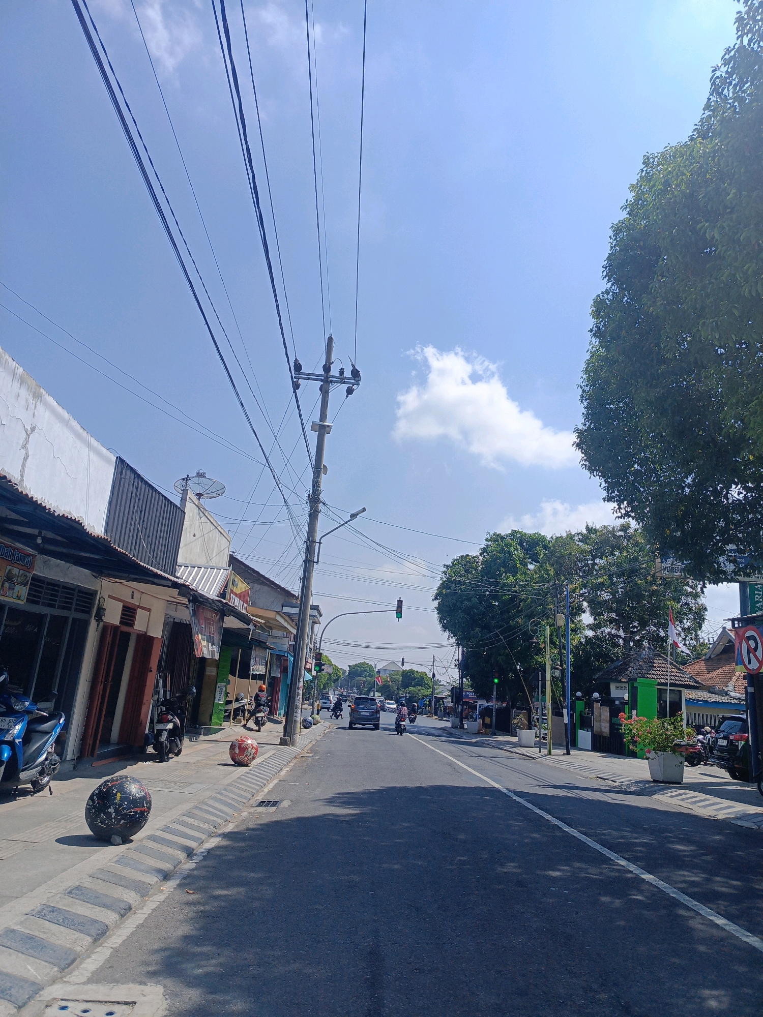 The image captures a street scene in Sukowinangun, Indonesia, on a clear, sunny day with a bright blue sky and scattered white clouds. The perspective is from a road looking forward.
On the left side of the street, there is a row of buildings that appear to be shops or small businesses, with awnings extending over the sidewalk. Some of these buildings have weathered white facades and metal roofs. Several motorcycles are parked along the sidewalk, including a blue scooter in the foreground. Two large, spherical decorative objects are placed on the sidewalk, one black with white and red splatters, and another red and white.
The road itself is paved with asphalt and has a white dividing line. Traffic is visible, including cars and motorcycles moving in both directions. A traffic light is visible in the distance, currently displaying a green light. A utility pole with numerous overhead wires stands prominently on the right side of the road, contributing to a network of crisscrossing cables across the sky.
On the right side of the road, there are more buildings, including one with a green facade and a tiled roof, and a prominent sign with a red circle and a diagonal line, indicating a prohibition. A large tree with dense green foliage overhangs the right side of the scene. The environment suggests a typical urban or semi-urban setting with commercial activity and active street life.