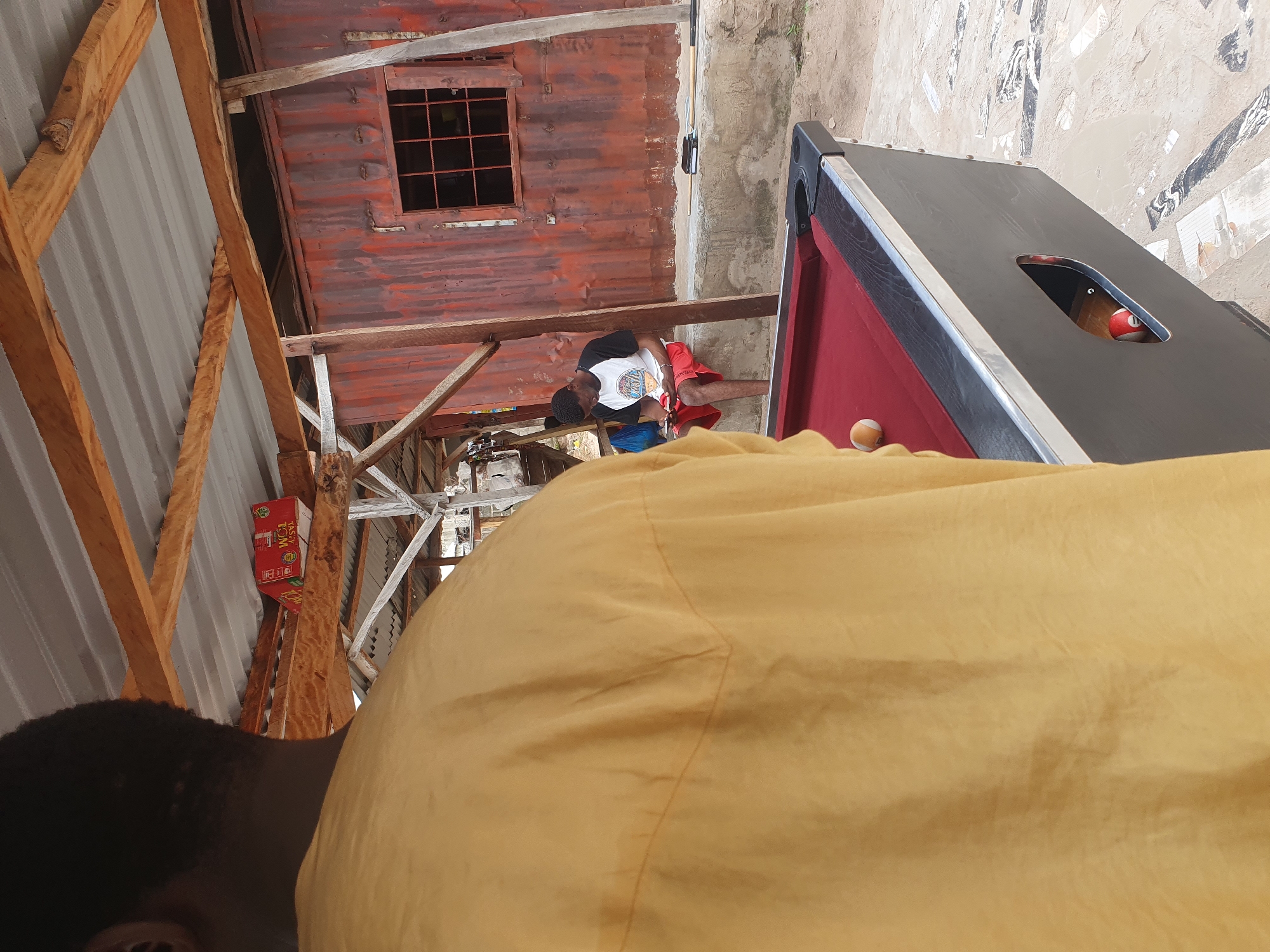The image depicts an outdoor leisure setting, likely a bar or a community gathering spot in Lokoja, Nigeria. In the foreground, a person from the back is wearing a yellow, long-sleeved shirt. A pool table with a red felt surface is visible, with a white and orange ball on it. A red and white pool ball is also seen inside the pocket of the table.
In the mid-ground, a young man is seated on a concrete ledge. He is wearing a black and white t-shirt with graphics and red shorts. He is looking down at his mobile phone, which he holds in his hands. A blue bag is behind him.
The environment appears rustic. A weathered, corrugated metal structure with a window featuring a grid is visible in the background. Wooden beams support a corrugated metal roof overhead. The ground is a mix of sand and what appears to be cracked concrete or paving stones. There is a red box with white lettering on one of the wooden beams. The overall impression is of a casual, open-air venue.