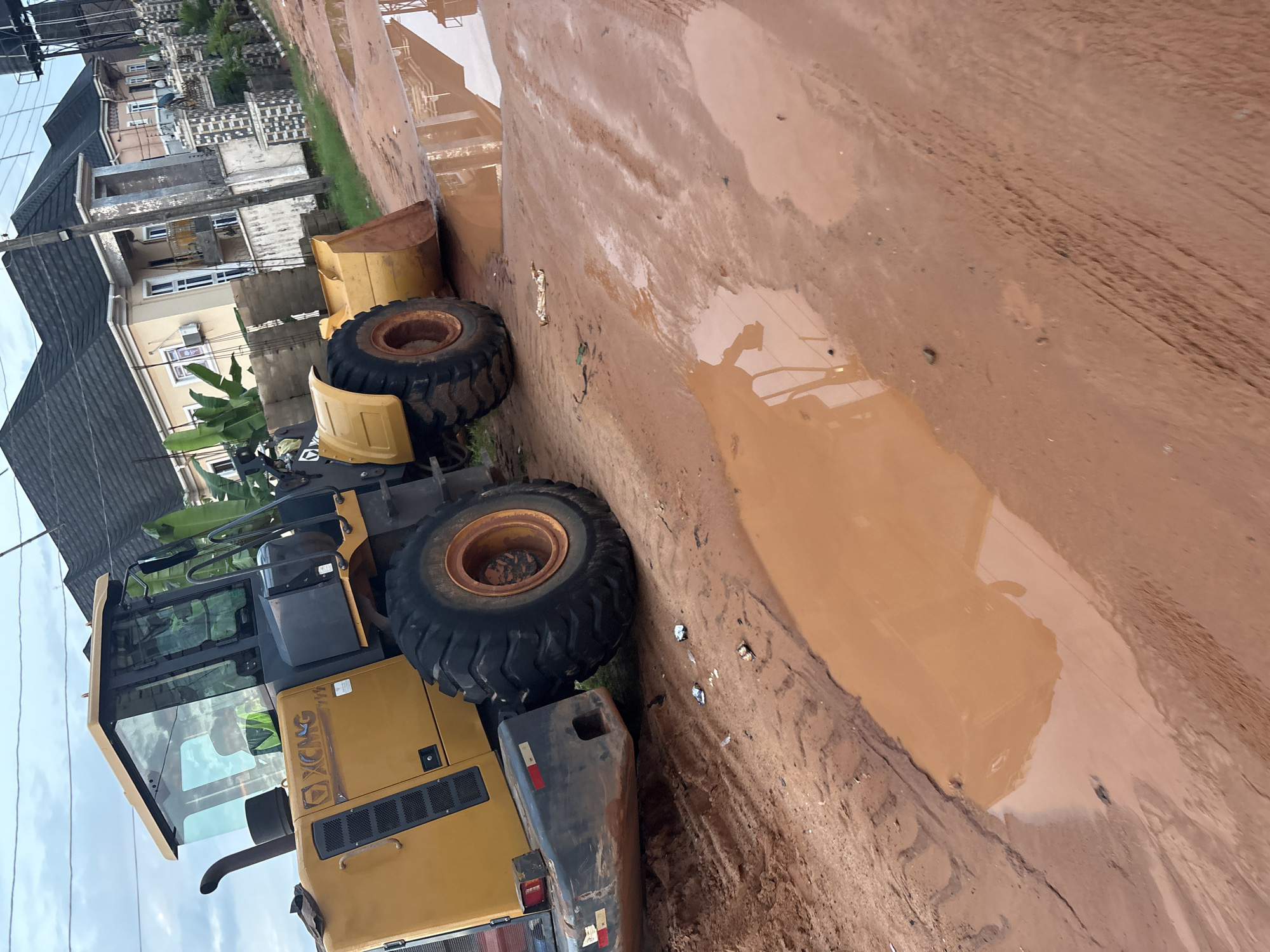 An elevated, medium shot captures a yellow XCMG wheel loader parked on a muddy, dirt road in Obosi, Nigeria. The loader occupies the left side of the frame, with its front bucket raised and resting on the ground. The tires of the loader are large and treaded, suggesting it's designed for heavy-duty work. The cab is visible, with a person wearing a high-visibility vest inside. The road is covered in reddish-brown mud and several large puddles, reflecting the overcast sky and surrounding buildings.
In the background, several modern houses with dark tiled roofs are visible. These houses appear to be multi-story dwellings, with some featuring balconies and ornate architectural details. The environment suggests a residential area undergoing construction or experiencing a period of heavy rain, leading to the muddy conditions. The overall scene depicts a common sight in areas with unpaved roads and significant rainfall.