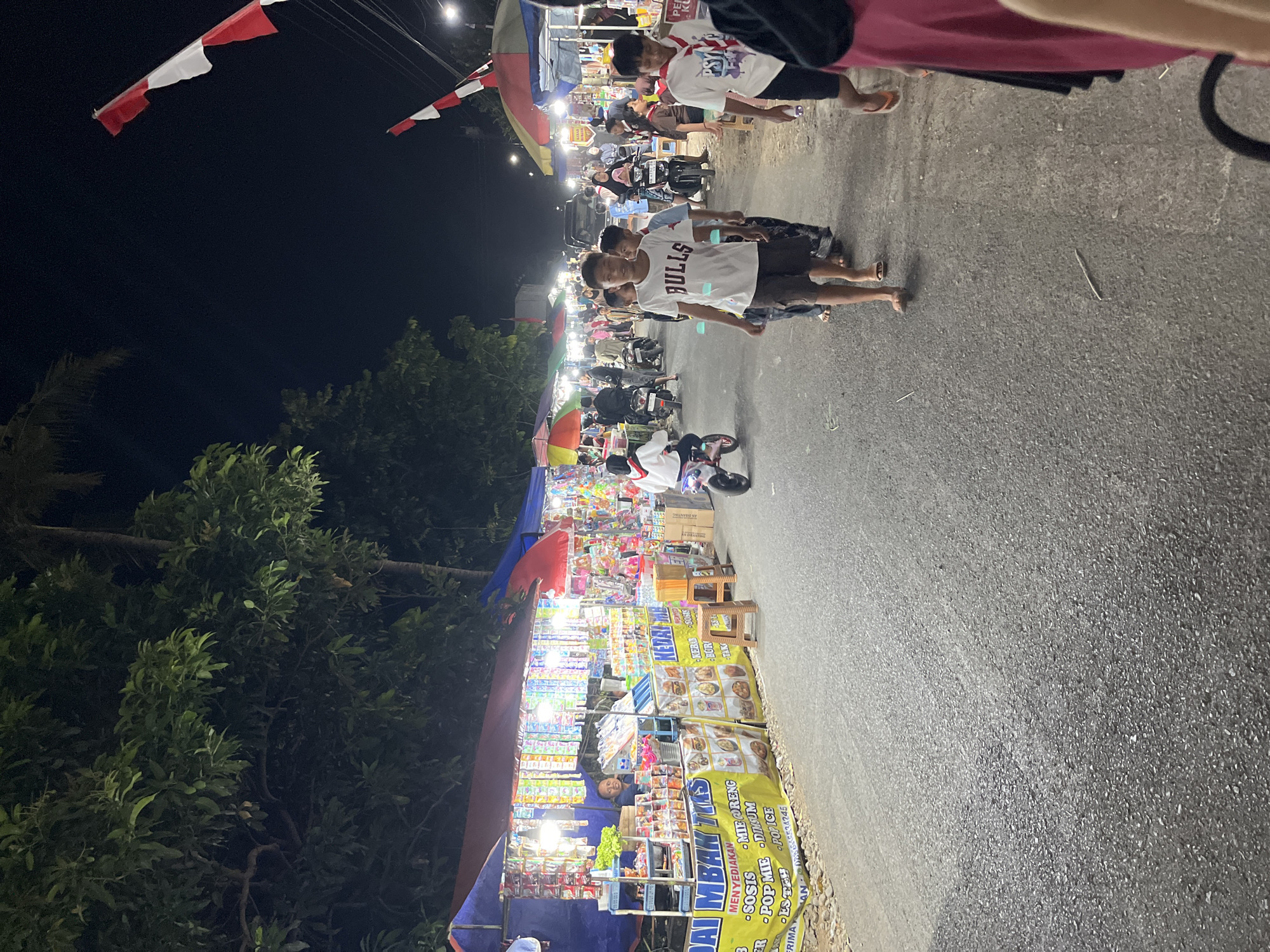 The image depicts a street market scene at night in Gembong, Indonesia. Numerous stalls line the road, adorned with colorful awnings and illuminated by string lights, selling a variety of goods, including snacks and food items. People of various ages are seen walking along the street, browsing the stalls, and interacting with each other. Some individuals are on motorcycles, navigating through the crowd. The atmosphere suggests a lively and bustling local event. The red and white Indonesian flags are visible, indicating a national celebration or event. The road surface appears to be asphalt, and trees are present on the sides of the street. The overall scene is one of a vibrant night market with a strong sense of community engagement.