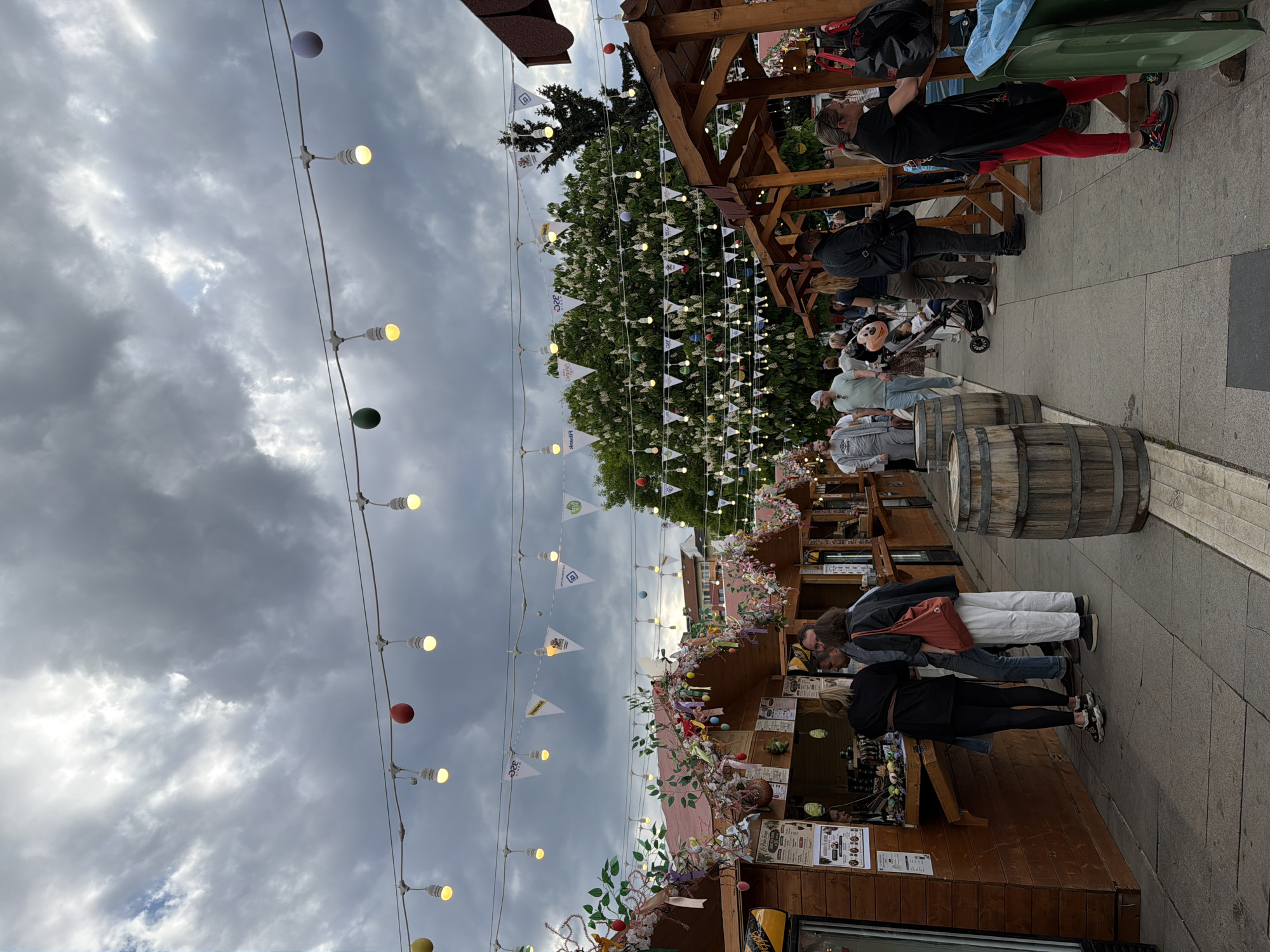 The image depicts a street market scene in Sofia, Bulgaria, under a cloudy sky. Several people are present, seemingly browsing the market stalls. The stalls are decorated with flowers, ribbons, and strung lights, and some have barrels placed in front of them. A series of decorative pennants are strung above the stalls, and there are decorative colored light bulbs hanging as well. The stalls appear to be constructed of wood, and their products are on display. The environment consists of a paved street.