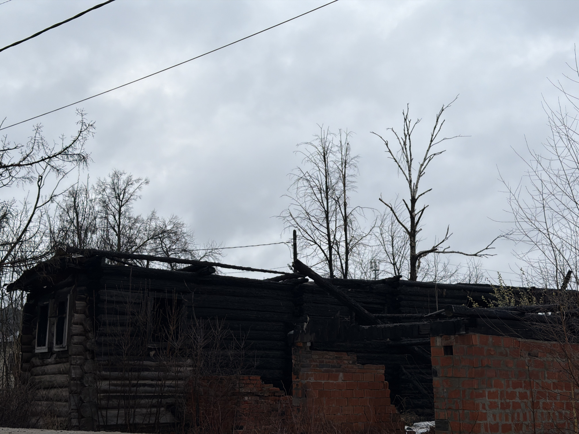 The image depicts the aftermath of a fire at a building in what appears to be a rural or semi-rural setting. The structure is a combination of charred logs and brickwork, indicating both the original construction materials and the extent of the damage. A portion of the building is reduced to rubble. The wooden parts are blackened, suggesting severe burning. Windows are visible on one side, though partially obscured by surrounding vegetation. The background features bare trees, hinting at either late fall or early spring. Above, the sky is overcast, contributing to the somber atmosphere. Based on the prompt's context, the location is identified as Chaikovsky, Russia. No people are visible in the scene.