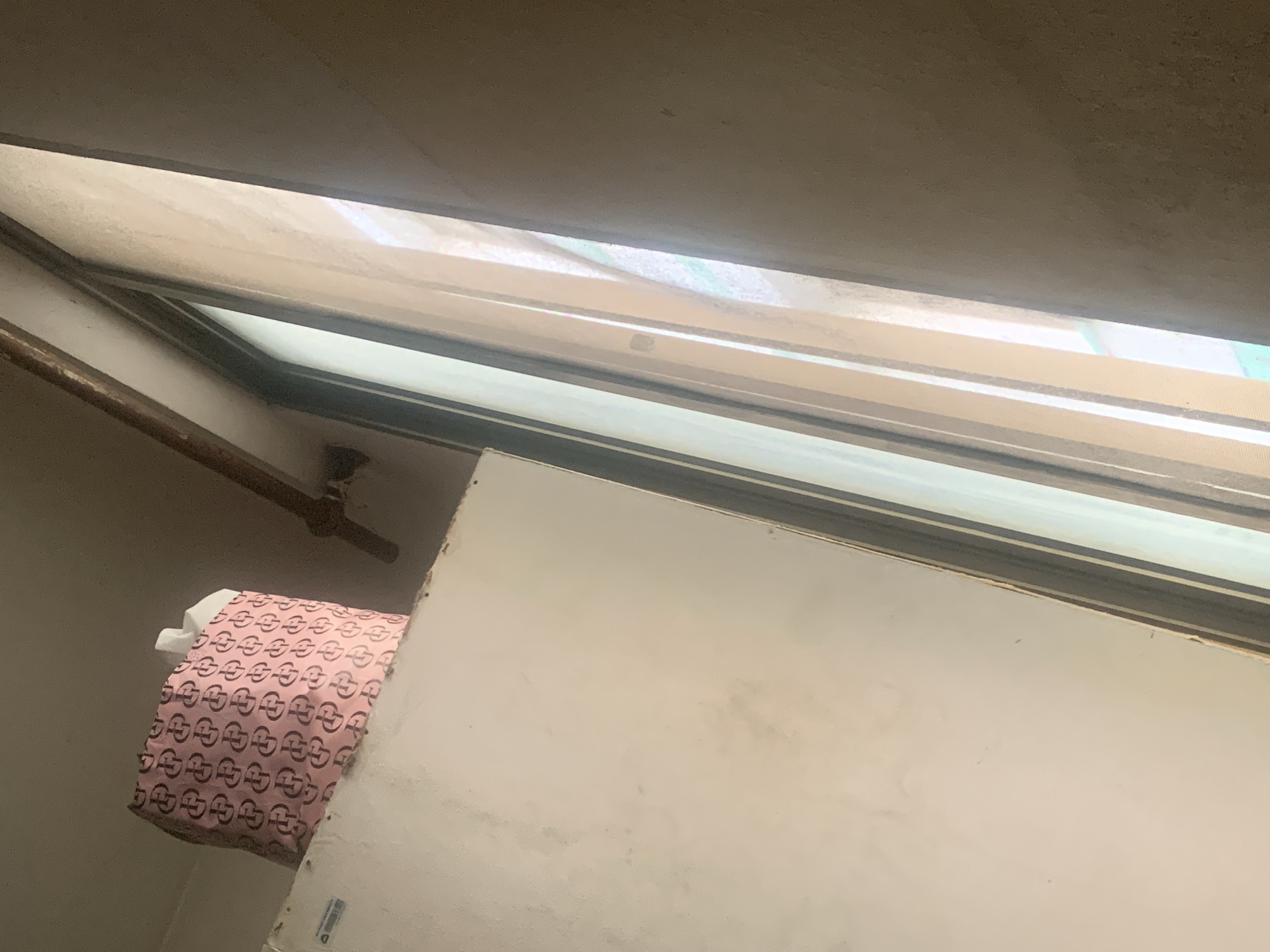 The photograph appears to be taken from inside a building in Dar es-Salaam, Tanzania, looking upward through a window. The window frame is visible, and there's a view of the outside, albeit obstructed by what appears to be a translucent covering. To the left of the window, a pipe runs horizontally along the wall. Below the window, a door or a panel is visible, and partially obscured by a pink patterned bag with a white object next to it. The scene seems to be indoors and dimly lit.