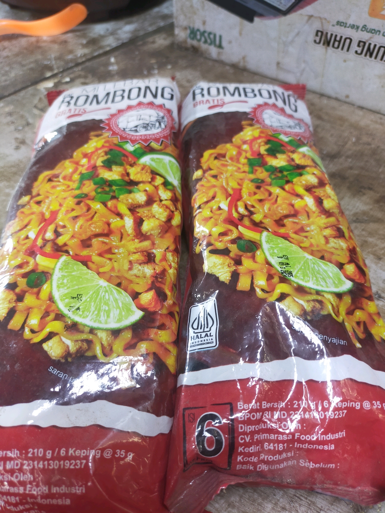 Two packages of "BOMBONG" brand instant noodles are shown from a slightly elevated, front-facing perspective. The packages are primarily red, with a large image of cooked noodles with lime wedges and green onions on each. The brand name "BOMBONG" is prominently displayed in white, capitalized letters at the top of each package. Below the brand name, "GRATIS" is written in smaller white letters. A red circular emblem with a line drawing of a vehicle is on the left package, and a HALAL logo is visible on the right package. The text on the bottom of the packages is partially obscured but indicates net weight, manufacturing details, and expiration information. One package has the text "saran penyajian" visible. The background consists of a rough, dusty surface and other packaging materials. The scene appears to be set indoors. No people are present in the image.