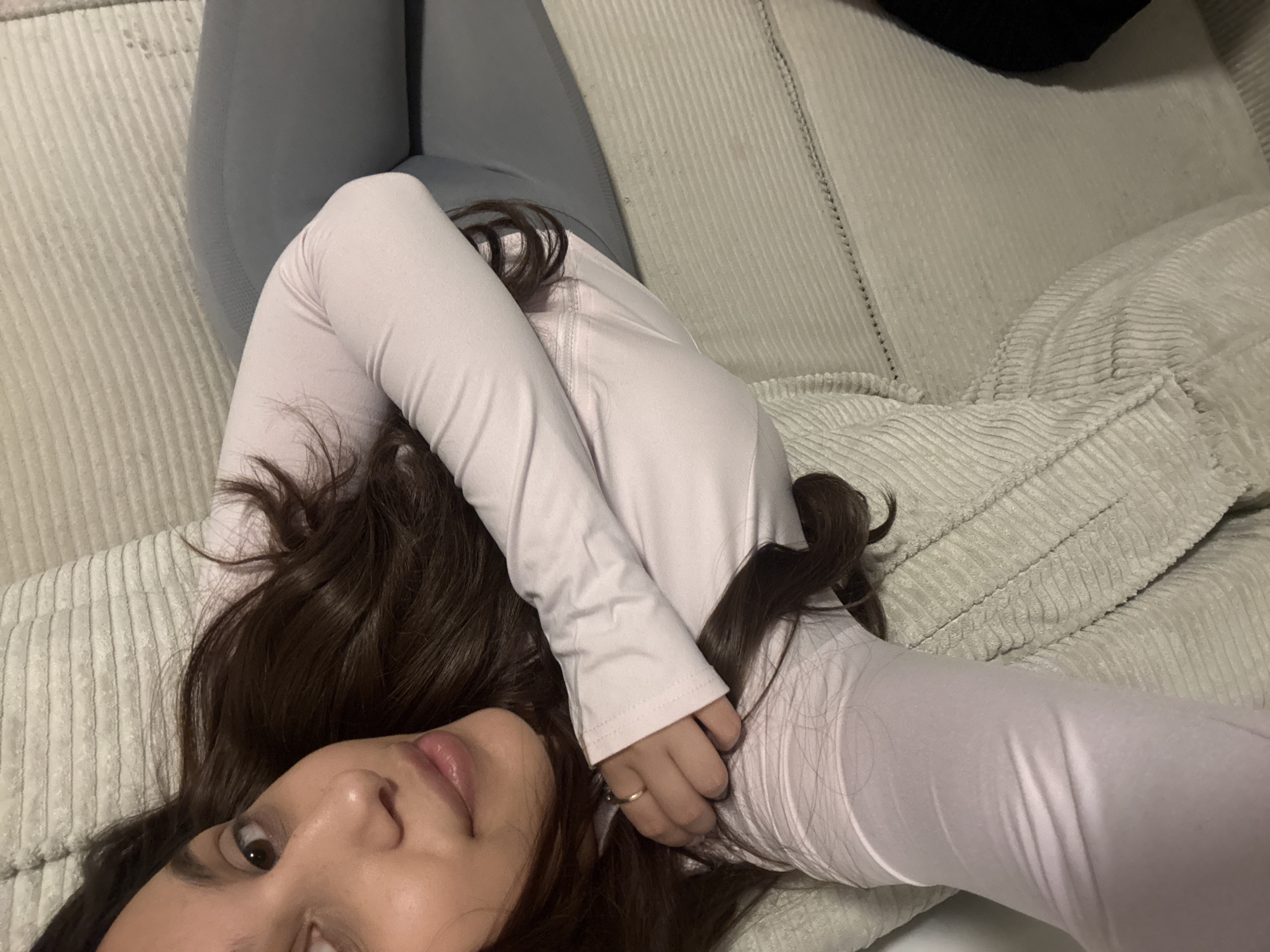 A woman with long brown hair lies on a light-colored corduroy couch. She wears light gray leggings and a light pink long-sleeved shirt. Her hands are intertwined near her head. A ring is visible on one of her fingers. The background shows the corduroy texture of the couch. The image does not provide information regarding the city of Mönchengladbach, Germany.