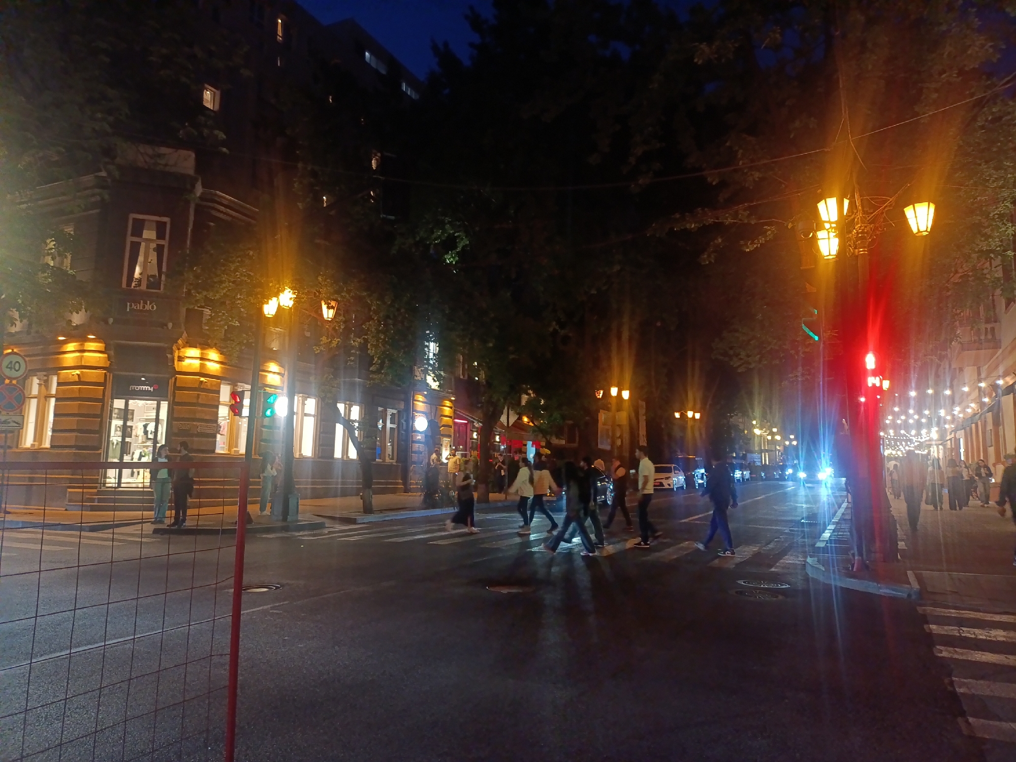 At night, the scene captures a street in Yerevan, Armenia. The street is busy with pedestrians using a crosswalk to traverse the road. The traffic signal displays a red light on one side and a green light on the other. The buildings on either side of the street are lit up, with storefronts visible. On the left, a building with warm lighting houses a store with mannequins in the windows. Street lights are visible and cast a warm glow. A metal barrier stands near the camera, marking a possible construction site. The sky is dark. Cars are also visible in the street.