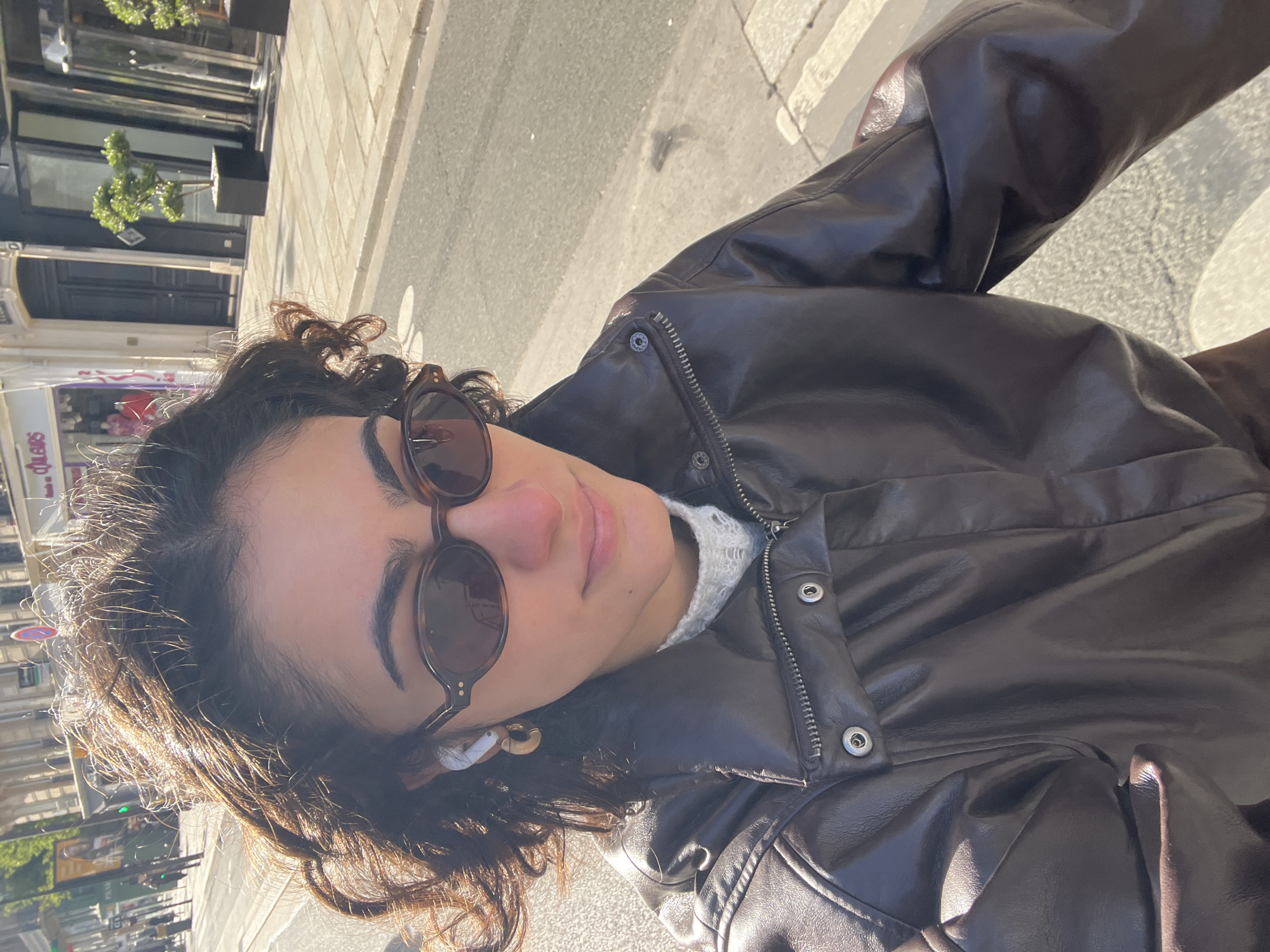 A medium-angle, candid eye-level shot depicts a woman wearing sunglasses and a brown leather jacket. She is walking on a street and appears to be taking a selfie. In the background, buildings, trees, and a crosswalk can be seen. The image suggests a sunny day in an urban environment, possibly in Paris, France.
