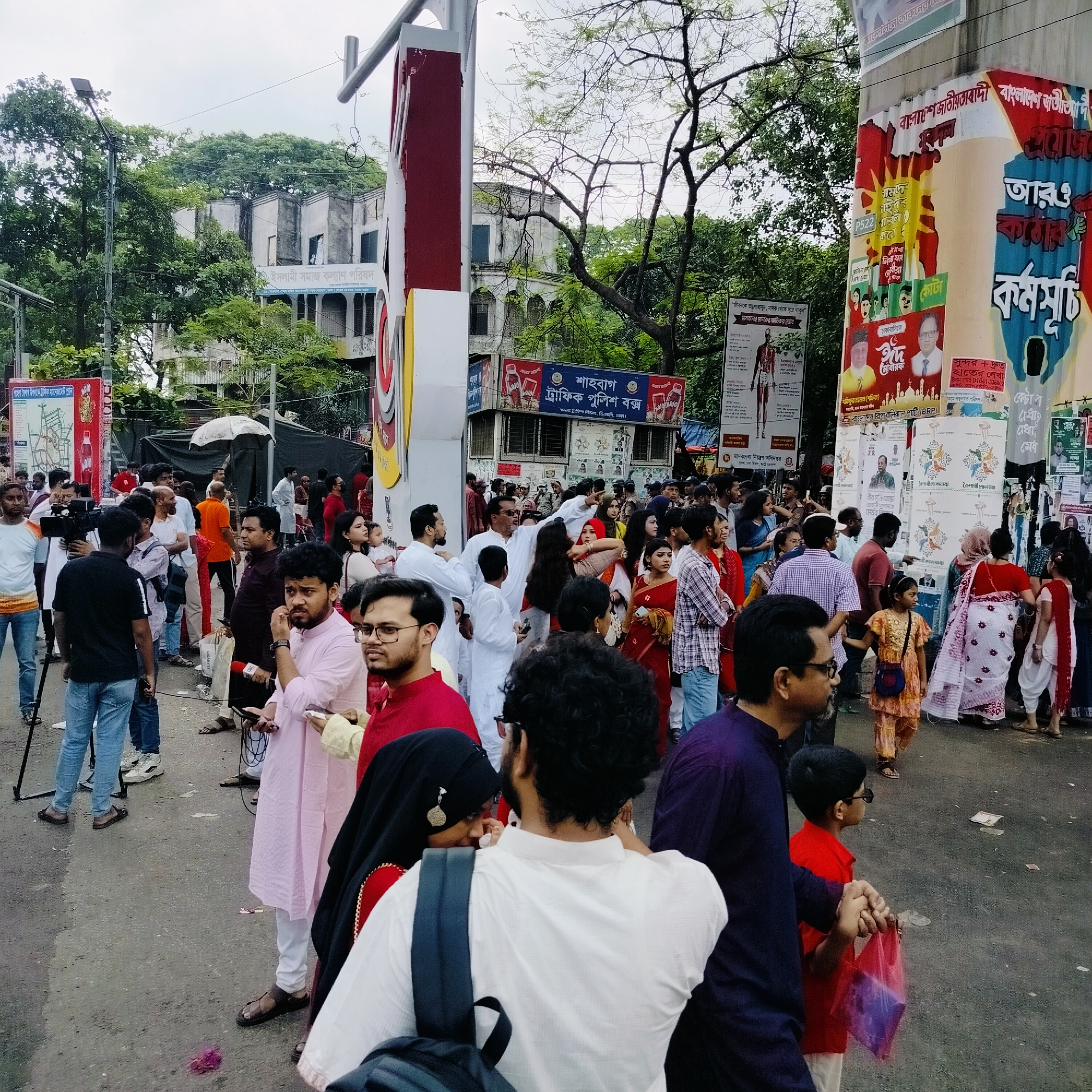This is a street scene in Dhaka, Bangladesh. It is daytime, and a large group of people, mostly wearing colorful traditional clothing, are gathered. Many are dressed in red, white, and pink clothing. Some individuals are wearing religious headdresses.
A person holding a camera on a tripod is filming. Several people are standing near the camera. Many posters with writing in Bengali are attached to nearby buildings and structures. Signs identify the location as Shahbag Traffic Police Box. A building with the name "Islami Samaj Kalyan Porishod" (Islamic Social Welfare Council) is visible in the background. Another structure, also covered with posters, is labeled as relating to "Pohila Boishakh" (Bengali New Year) traffic management.
