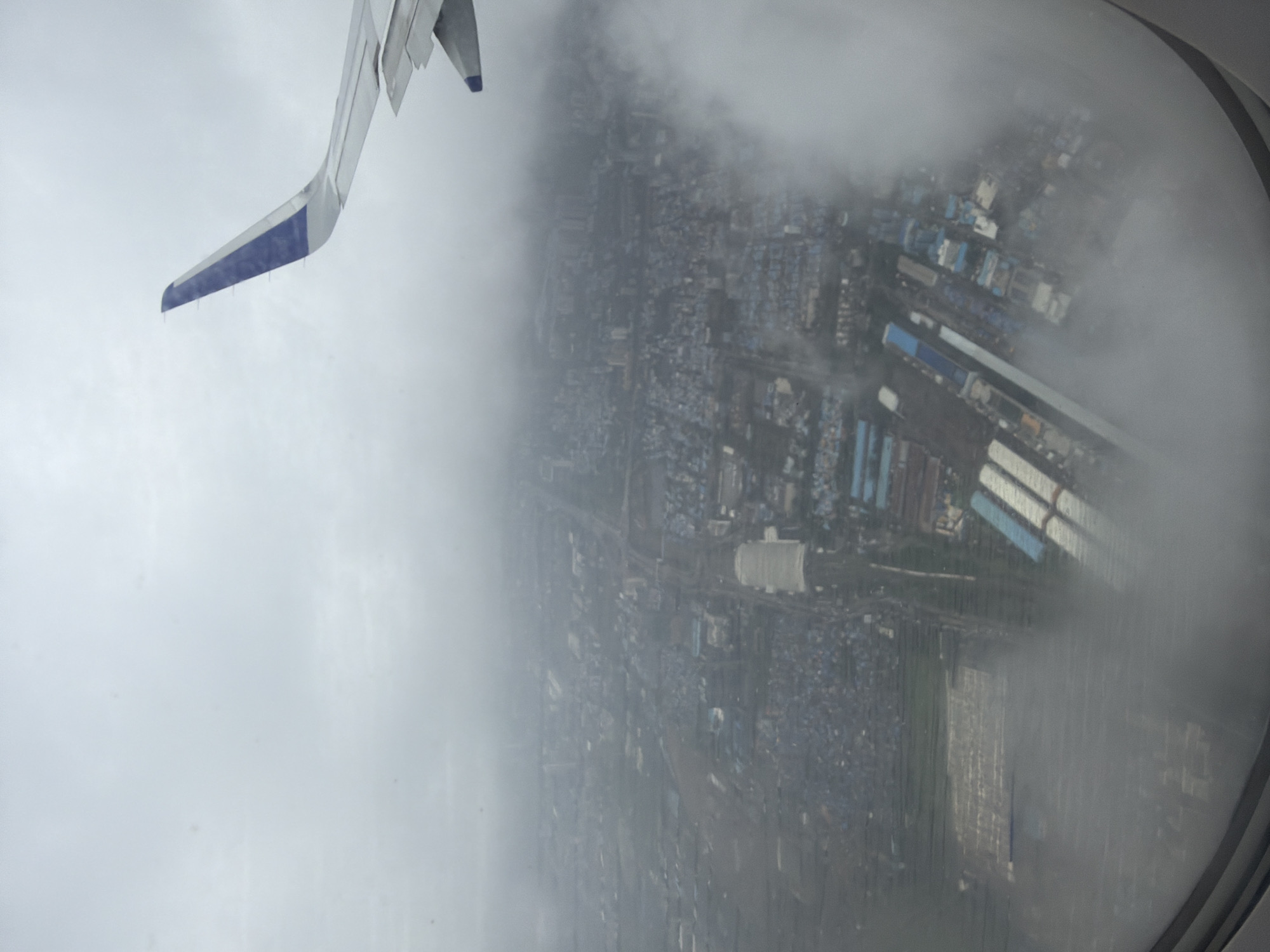 The image is an aerial view from an airplane window, looking down at a dense urban and industrial area, possibly Navi Mumbai, India, under a cloudy sky. An airplane wing, with a blue and white tip, is visible in the upper right corner. The landscape below is characterized by a mix of residential buildings with blue roofs, industrial structures, and some open, possibly agricultural land in the lower left. A large, elongated building with a white roof appears to be a sports stadium or a large industrial complex in the middle left of the frame. Roads and railway lines weave through the city. Clouds partially obscure the view of the ground, creating a hazy and diffused atmosphere. The image depicts a sprawling cityscape under overcast conditions, seen from a high altitude.