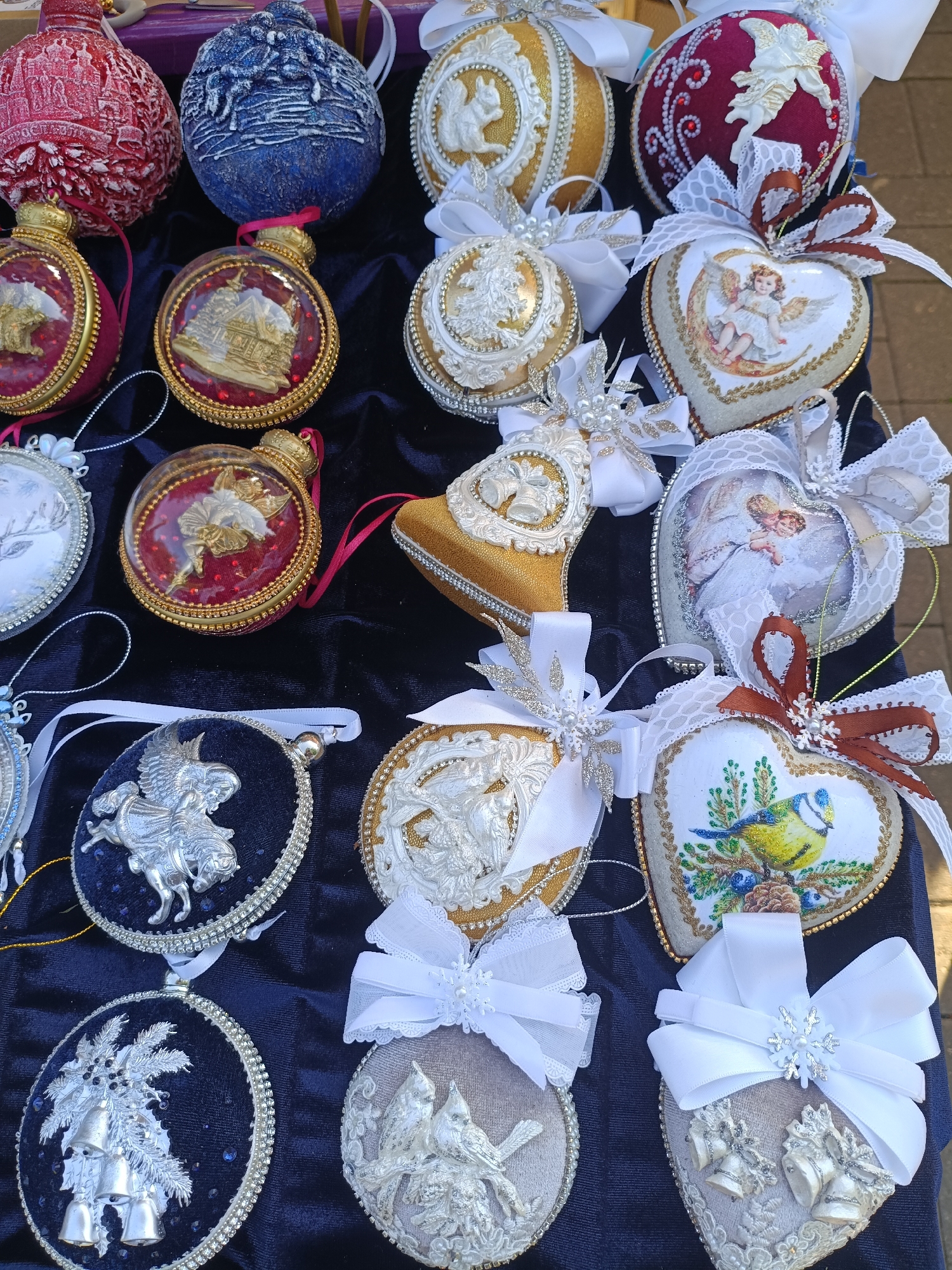 The image displays a collection of handcrafted Christmas ornaments arranged on a dark blue velvet cloth. The ornaments are varied in shape and design, with many featuring elaborate embellishments, glitter, and ribbons. Several of the ornaments are spherical, while others are heart-shaped or oval. Decorative elements include religious figures, animals, and floral motifs. The setting appears to be an outdoor market stall, suggested by the visible paving stones in the background. There are no people visible in the image. The lighting is natural, indicating it was taken during the daytime. The overall impression is one of festive, artisanal holiday decorations.
