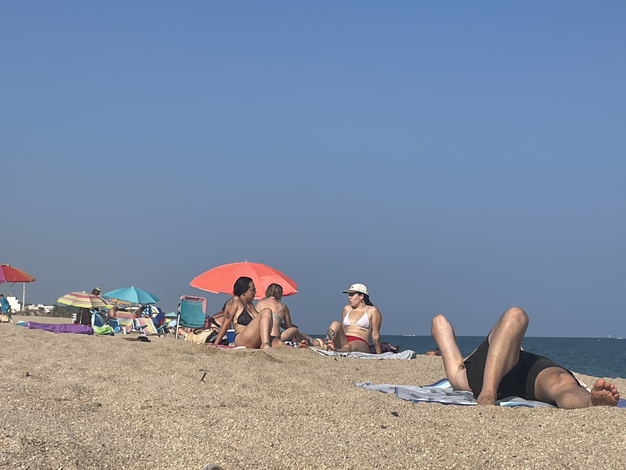 The image depicts a sunny day at the beach in Canet de Mar, Spain. The foreground is dominated by a sandy beach with small pebbles. In the mid-ground, several people are visible enjoying the sun. Three women are seated together under a large orange umbrella. One woman is wearing a black bikini, another a light-colored bikini, and the third a white bikini and a light-colored hat. Further to the right, a person is lying on their back on a towel, wearing dark green swim trunks. In the background, the blue ocean stretches to the horizon under a clear blue sky. Scattered beach umbrellas and chairs are present, along with distant buildings and structures suggesting a coastal town.