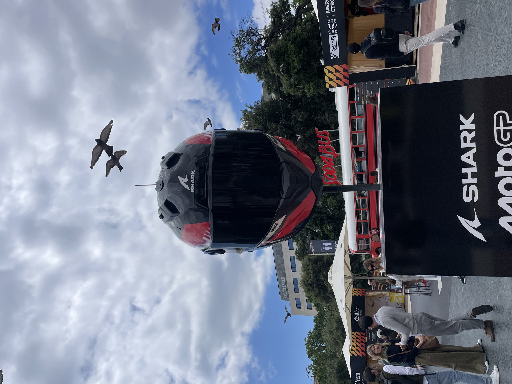 A large black and red Shark motorcycle helmet is suspended in mid-air, several pigeons are flying around it. Below, there is a red and white "Sodabus" double-decker bus. There are crowds of people around the bus. The image appears to be taken in an urban setting, possibly Barcelona, Spain, based on the "Circuit de Barcelona Catalunya" sign. There is a white building in the background. A sign with the Shark Moto GP logo is visible, too.