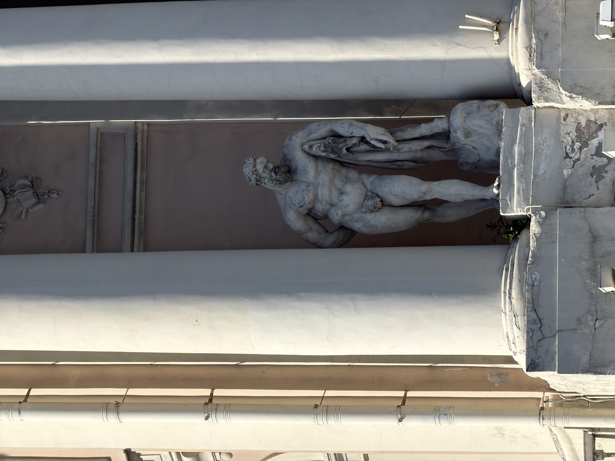This image presents a large, classical-style statue of a male figure situated between two substantial architectural columns. The statue, depicting a muscular, bearded man in a dynamic, semi-reclining pose with drapery flowing around his left arm and shoulder, is made of a light-colored stone and exhibits signs of weathering and age with visible cracks and worn surfaces. He stands on a pedestal, which is also aged and chipped, integrated into the building's facade.
The two flanking columns are tall, white, and fluted, with their bases showing significant deterioration, including crumbling material and dark streaks. Behind the statue, the building's wall is a muted, light brownish-pink color. On the left side of this wall, subtle decorative architectural elements or carvings are discernible.
The scene is well-lit by direct sunlight, indicating a daytime setting, likely morning or afternoon, which casts strong shadows and highlights the textures of the stone and the weathered surfaces. The architectural style, featuring classical columns and a large figurative sculpture, suggests a grand, historic building, consistent with the urban landscape of Saint Petersburg, Russia. There is no visible text in the image.