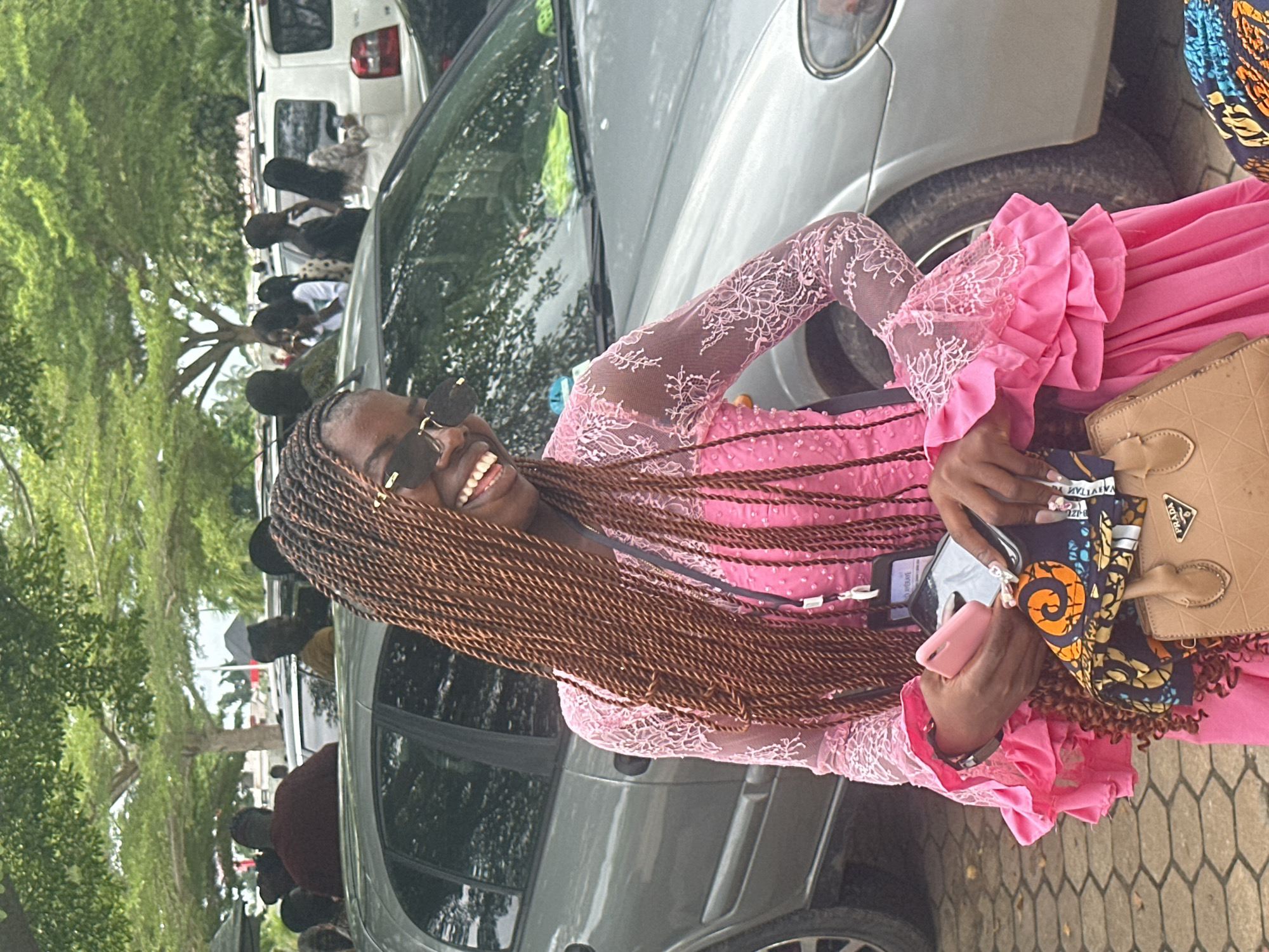 In Itoga, Nigeria, a young Black woman with long, dark braided hair and sunglasses is posed in front of a silver car. She is wearing a bright pink dress with lace detailing on the sleeves and a ruffled hem. In her hands, she holds a pink phone and a small brown bag with a Prada logo. Behind her, other people and vehicles are visible, suggesting a public gathering or event. The ground appears to be paved with hexagonal tiles, and trees provide shade overhead.