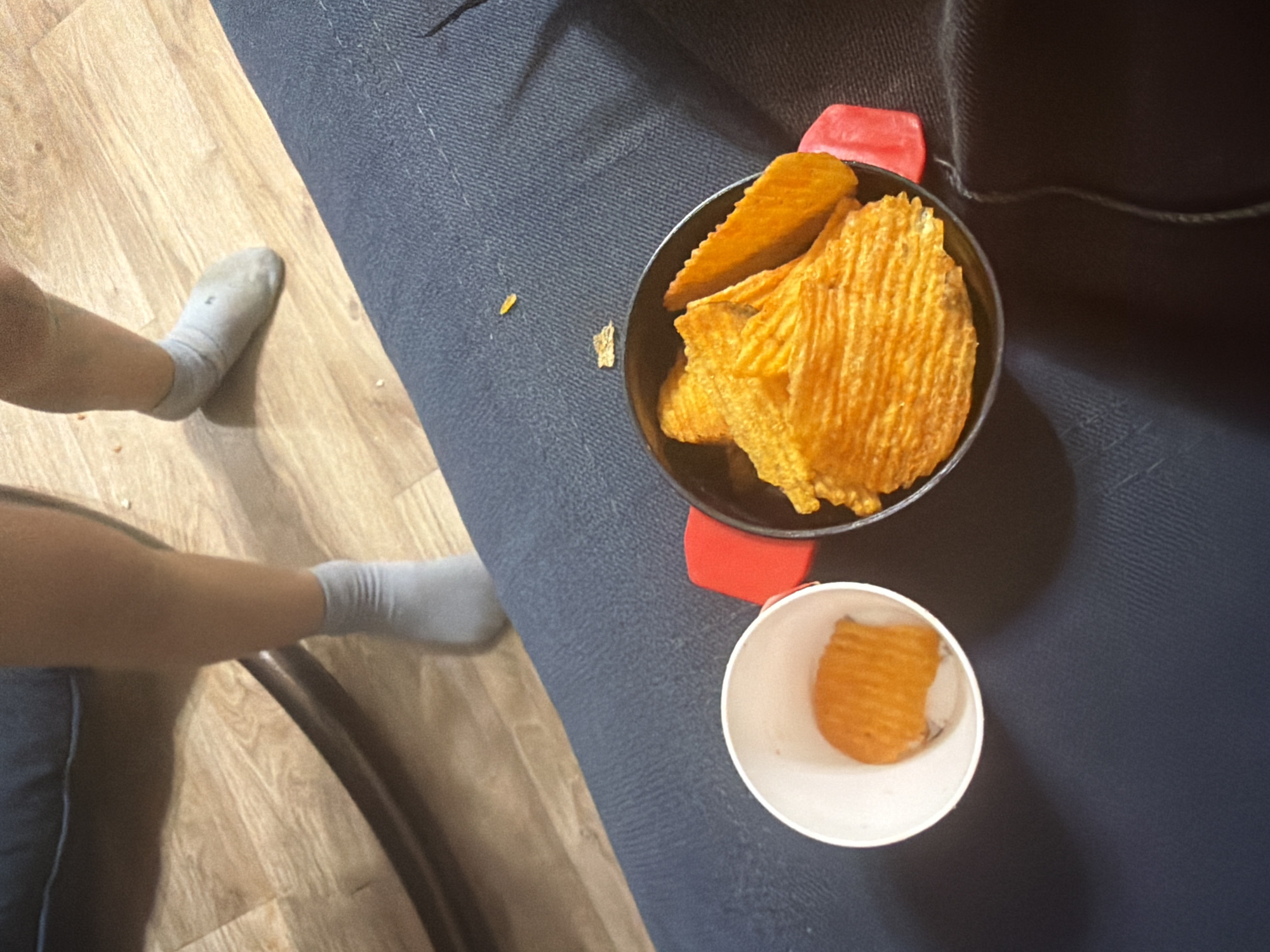An overhead shot shows two servings of ridged potato chips. One serving is in a small white paper cup, with a single chip visible. The other serving is in a black bowl, filled with multiple chips. A person's legs, wearing light grey socks, are visible in the upper half of the image, standing on a light wood-grain floor. The background is a dark blue upholstered surface, possibly a couch. The image appears to be taken indoors, with no specific landmarks to indicate a location in Shymkent, Kazakhstan.