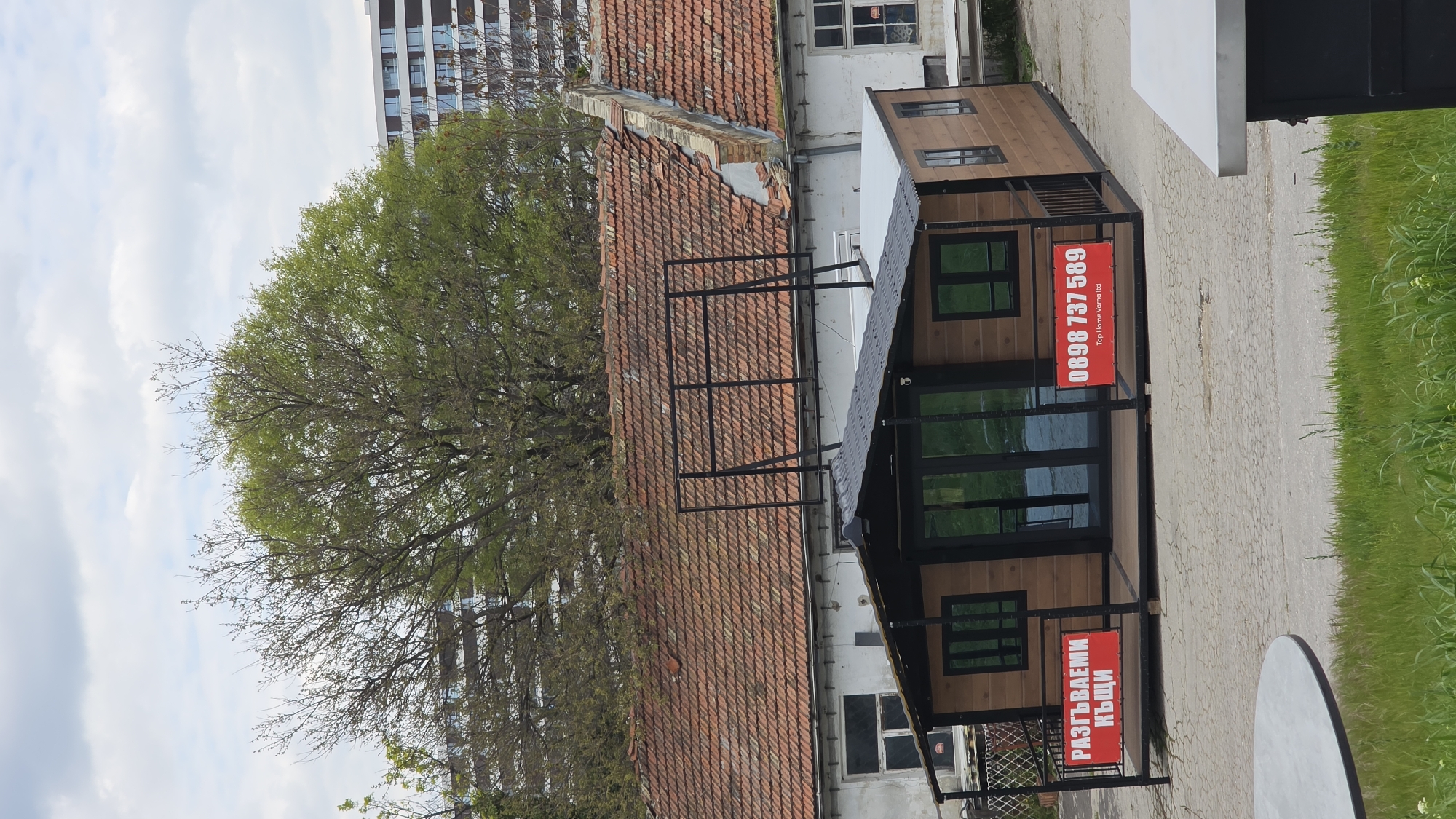 The image depicts a small, modern-style building in Varna, Bulgaria. The structure has a wooden facade with black frames around the windows and a sign reading "РАЗГЪВАЕМИ КЪЩИ" in Bulgarian. Another sign displays the phone number "0898 737 589" and the name "Top Home Varna ltd."
The building is situated next to an older structure with a traditional red tile roof. A large tree with green foliage partially obscures the buildings and provides a backdrop. A taller modern building with many windows rises in the background. The ground appears to be paved, with some grass and greenery visible on the periphery. The sky is cloudy.