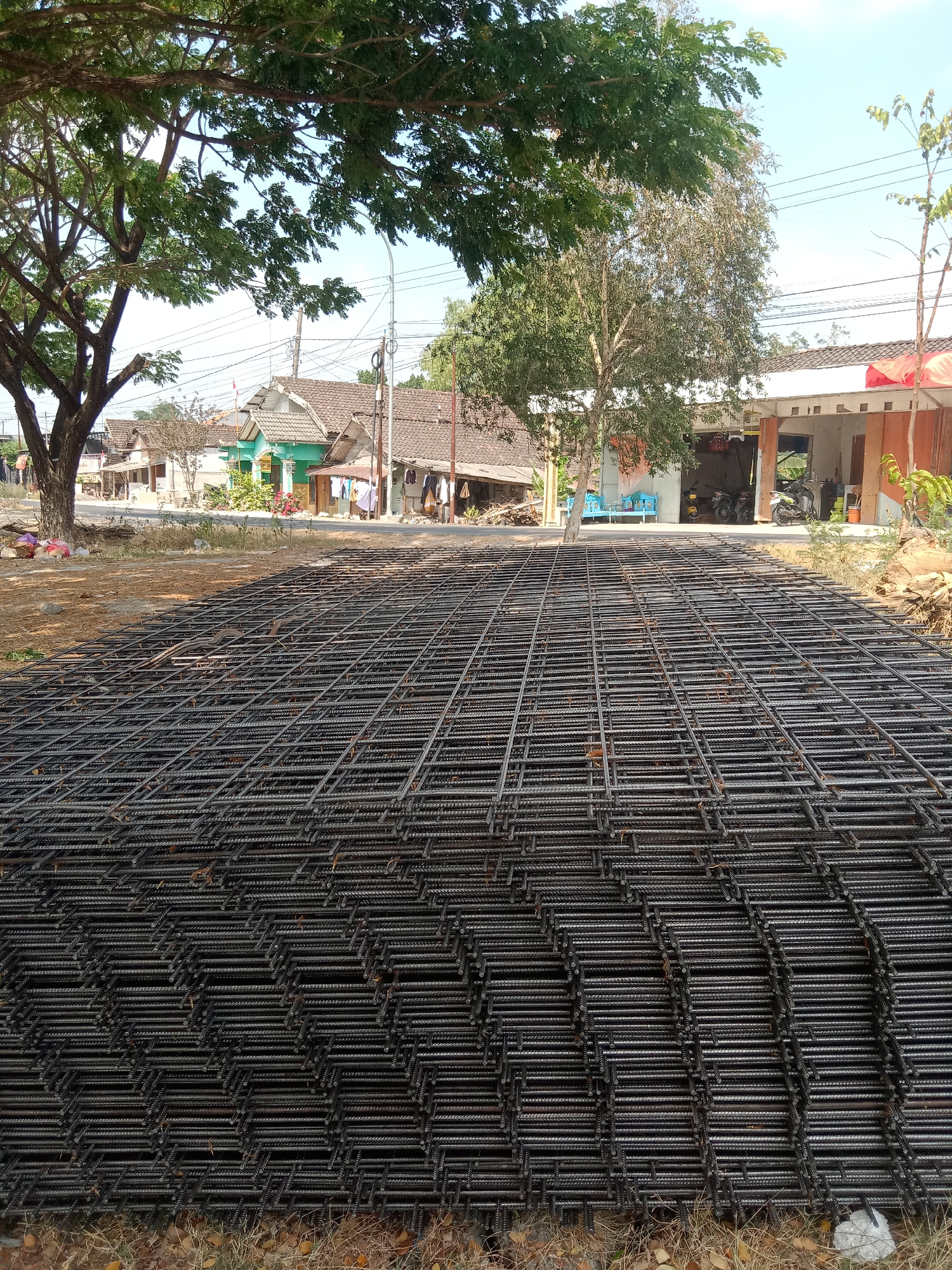 In the foreground, a large stack of rebar mesh, used in construction, lies on a dry, dusty ground. The rebar is dark gray and forms a grid pattern. Behind the rebar, a road runs horizontally, with houses and trees on the other side. The houses have tiled roofs and appear to be of modest construction. Some laundry is hanging outside one of the houses. Further back, power lines stretch across the sky. The overall scene suggests a semi-urban or rural environment in Siwalan, Indonesia, possibly a construction site or a storage area for building materials. The weather appears to be sunny, with a clear blue sky.