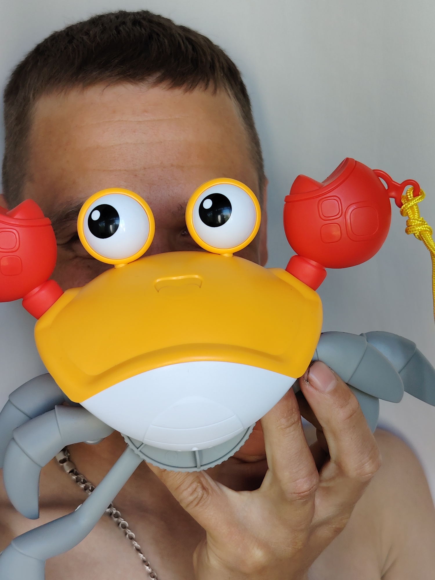 A man is holding a yellow, white, red, and grey plastic toy crab in front of his face. His head is visible above the toy, and his hand is holding the crab. A silver chain is visible around his neck. The background is a plain white wall. The location is Chaikovsky, Russia.