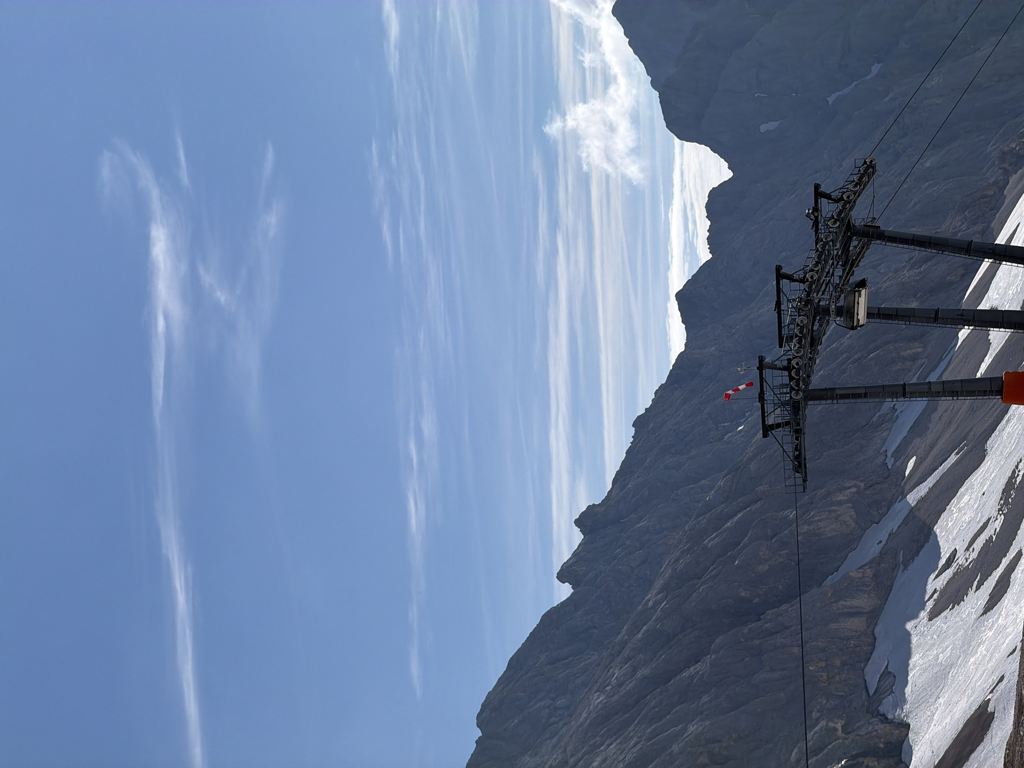 The image shows a cable car station on a mountainous landscape. The station has a grey metal structure with multiple pulleys and a cable extending from it. A red and white striped flag is mounted on the station. The mountains in the background are steep and rocky, with patches of snow on their slopes. The sky is blue with wispy white clouds. This scene is likely at the Zugspitze, Germany's highest peak, located near Garmisch-Partenkirchen. There are no people visible in the image.