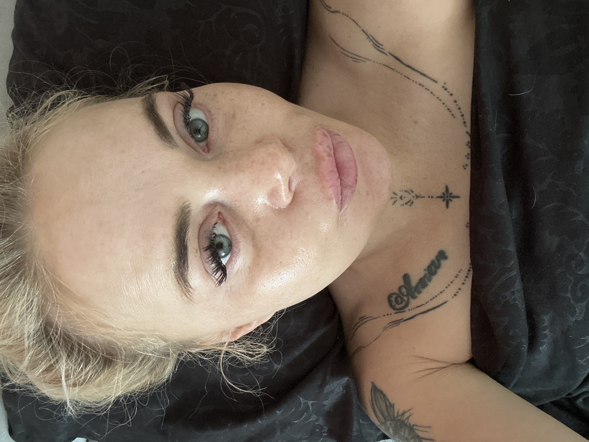 A close-up, overhead shot depicts an adult female lying on a dark, textured surface, likely bedding. Her blonde hair is loosely pulled back. She has green-blue eyes, dark eyebrows, and long eyelashes, gazing directly at the camera. Her skin exhibits freckles on the nose and cheeks, with a noticeable sheen on her forehead, around the eyes, and on the nose and upper lip. Her lips are full and pouty, appearing reddish-pink. Visible tattoos include a dark, partially visible design on her right upper arm/shoulder, the word "Amore" in stylized script with decorative elements on her right collarbone area, a geometric design with a star element on her lower neck/sternum, and linear abstract designs on her left collarbone. Her shoulders and chest are visible, covered by the dark material below the collarbone. The overall lighting is soft and even.