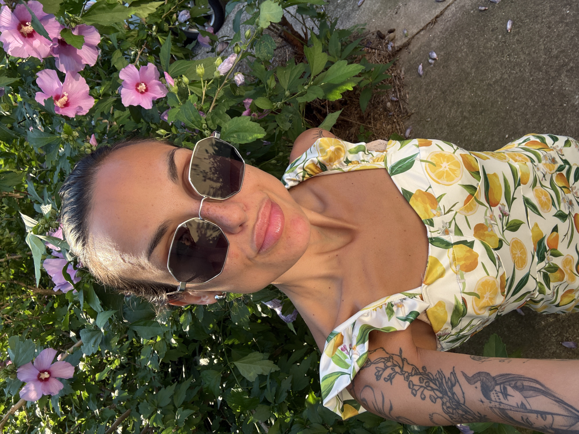 In this overhead, natural light selfie, a woman with dark hair pulled back and wearing large, hexagonal-shaped sunglasses is positioned in the center of the frame. She is outdoors, surrounded by lush green foliage and several blooming pink hibiscus flowers. Her dress features a vibrant yellow lemon print. On her left arm, a tattoo of interwoven branches and a stylized horned figure is visible. The background consists of green leaves, pink flowers, and a concrete path with fallen petals scattered across it. The sunlight suggests it is daytime, likely a warm and pleasant day. The woman's pose and the bright floral surroundings evoke a sense of casual enjoyment and connection with nature. No text is visible in the image. The location context provided is Pleven, Bulgaria.