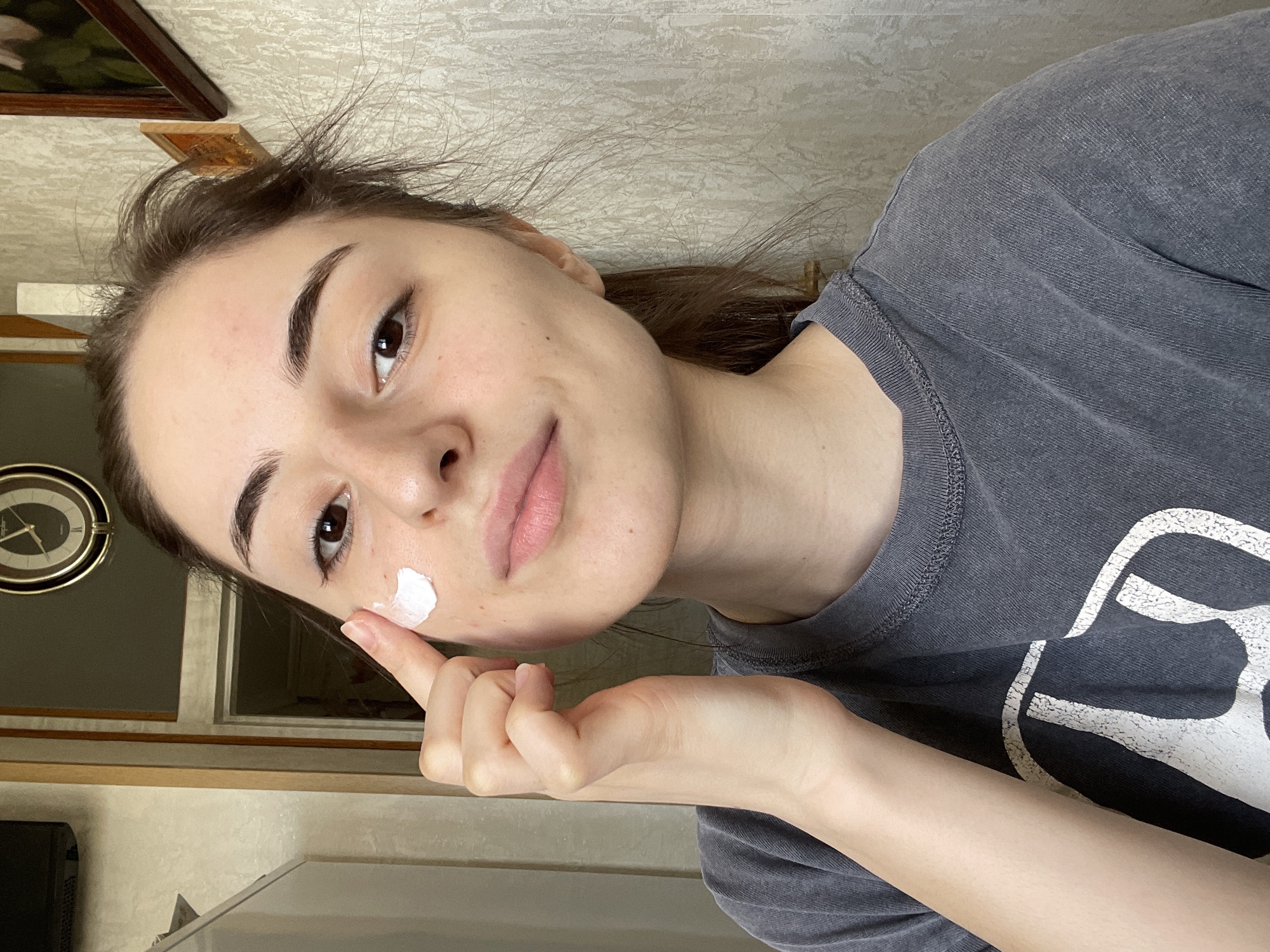 A young woman with dark hair and eyebrows is applying a dab of white cream to her cheek with her right index finger. She is wearing a gray t-shirt with a white graphic design. The background includes a patterned wall, a wooden framed painting, a wall-mounted clock, a window, and part of a cabinet. There is no definitive information in the image to confirm this scene is in Moscow, Russia.