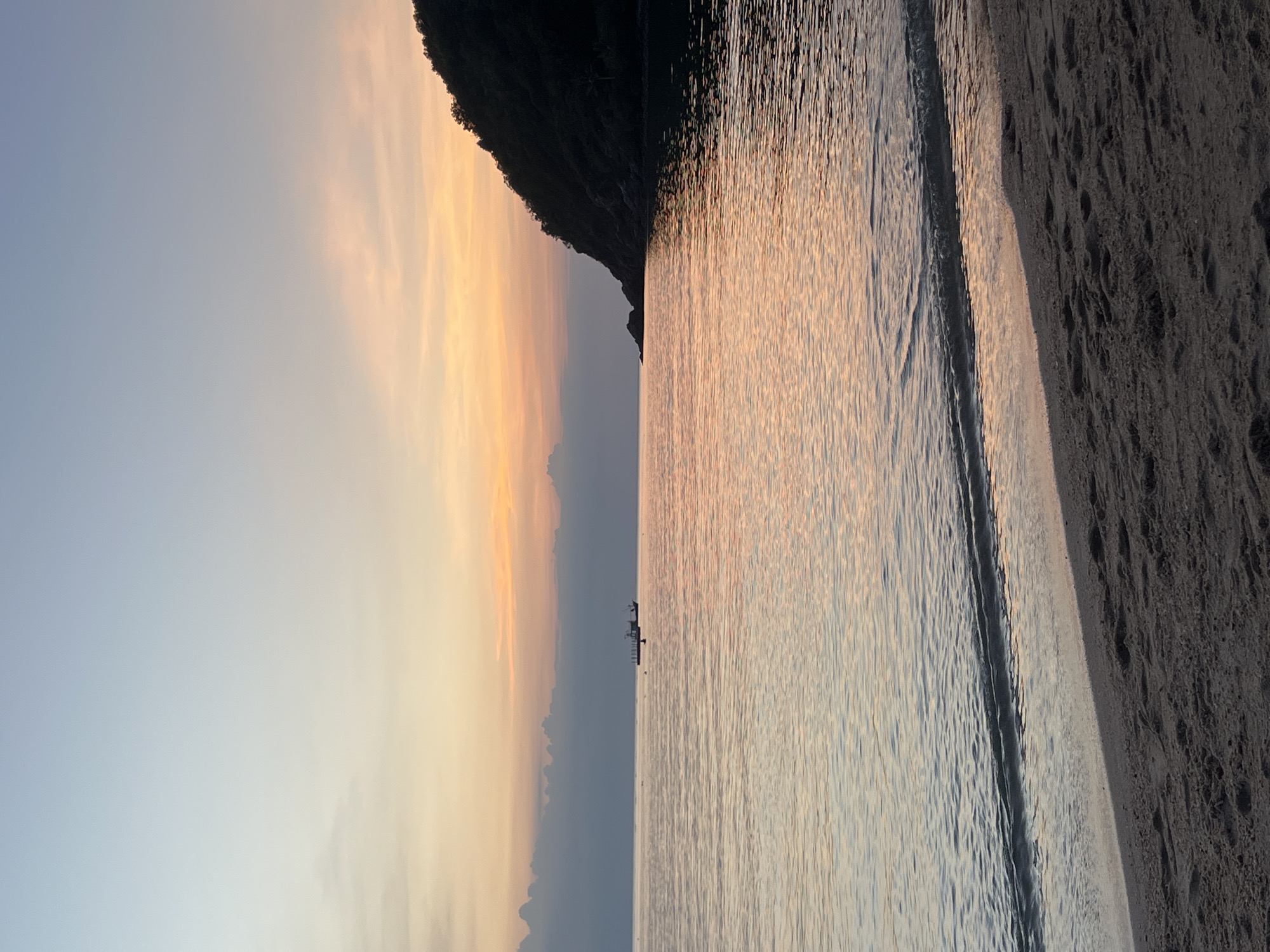 A serene beach scene at dusk in the Ko Pha-ngan District, Thailand. The sky displays a gradient of soft blues and warm orange and pink hues, indicative of sunset. The calm sea reflects the colors of the sky, with gentle waves lapping the sandy shore in the foreground. A dark, forested hill juts into the sea on the right side of the frame. In the middle distance, a small boat is anchored on the water, with a few figures visible on its deck. The beach itself is composed of dark sand with visible footprints. The overall atmosphere is tranquil and picturesque.