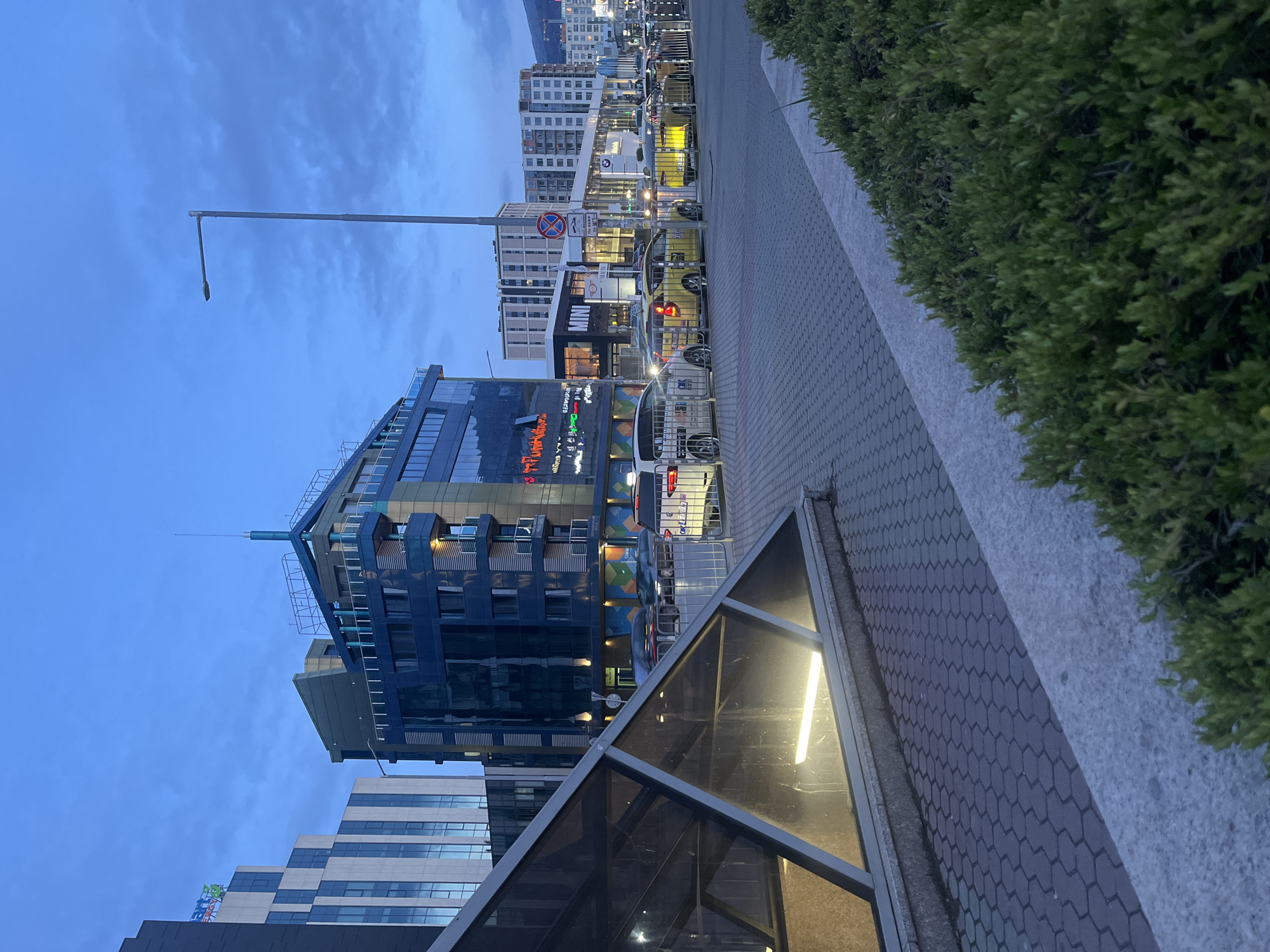 The image depicts a street view in Sofia, Bulgaria, at dusk. A modern, multi-story building with blue glass panels and a unique architectural design is prominently featured. The building's facade displays illuminated signs and artistic elements. A streetlight extends above the building.
Alongside the building, a paved walkway is visible, with hexagonal patterned paving stones and a concrete border. Adjacent to the walkway is a low, rectangular structure with a glass or transparent plastic covering, likely the entrance to an underground or partially submerged area. Beyond the walkway, a row of parked vehicles and other buildings can be seen. Dense green shrubbery lines the edge of the walkway. The sky is a muted blue, suggesting twilight or a cloudy day. The overall impression is of a contemporary urban landscape.