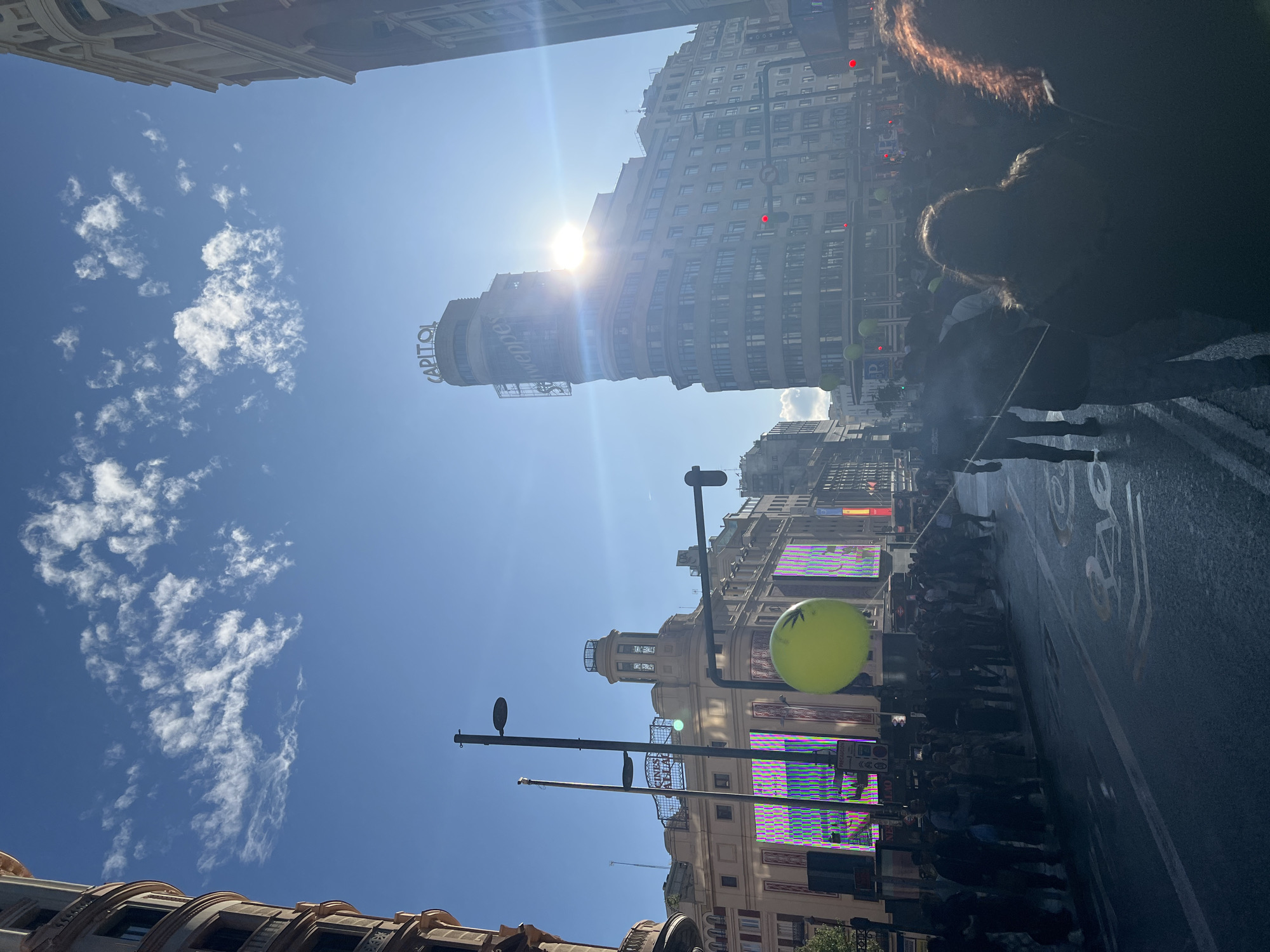 The image captures a street scene in Madrid, Spain. The iconic Carrión Building, often called the Capitol Building due to the illuminated "Capitol" sign, dominates the view. The sun is high in the sky, causing lens flare. A crowd of people is gathered on the street, with several green balloons visible. Streetlights are present in the foreground. The road markings are clearly visible. The sky is blue with scattered clouds.