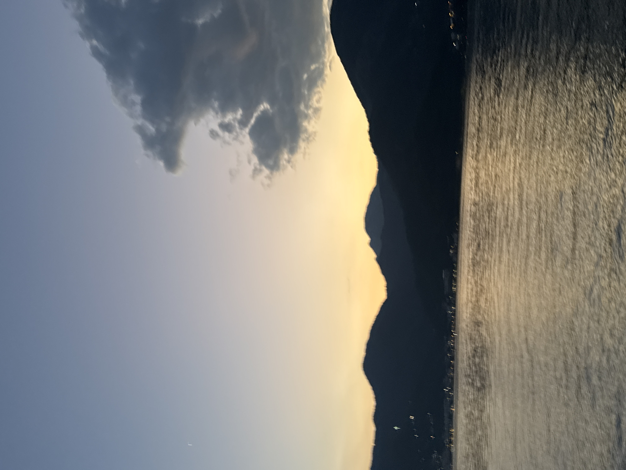 The image shows a serene landscape of mountains, a body of water, and a twilight sky. The mountains are silhouetted against a gradient sky that transitions from a pale blue at the top to a warm yellow-orange near the horizon, suggesting either sunrise or sunset. A large, dark cloud is visible in the upper right portion of the sky. The water in the foreground is rippling, reflecting the warm colors of the sky. Along the base of the mountains, distant lights are visible, indicating a settlement or town. There are no people or specific identifiable objects or actions depicted in the scene.