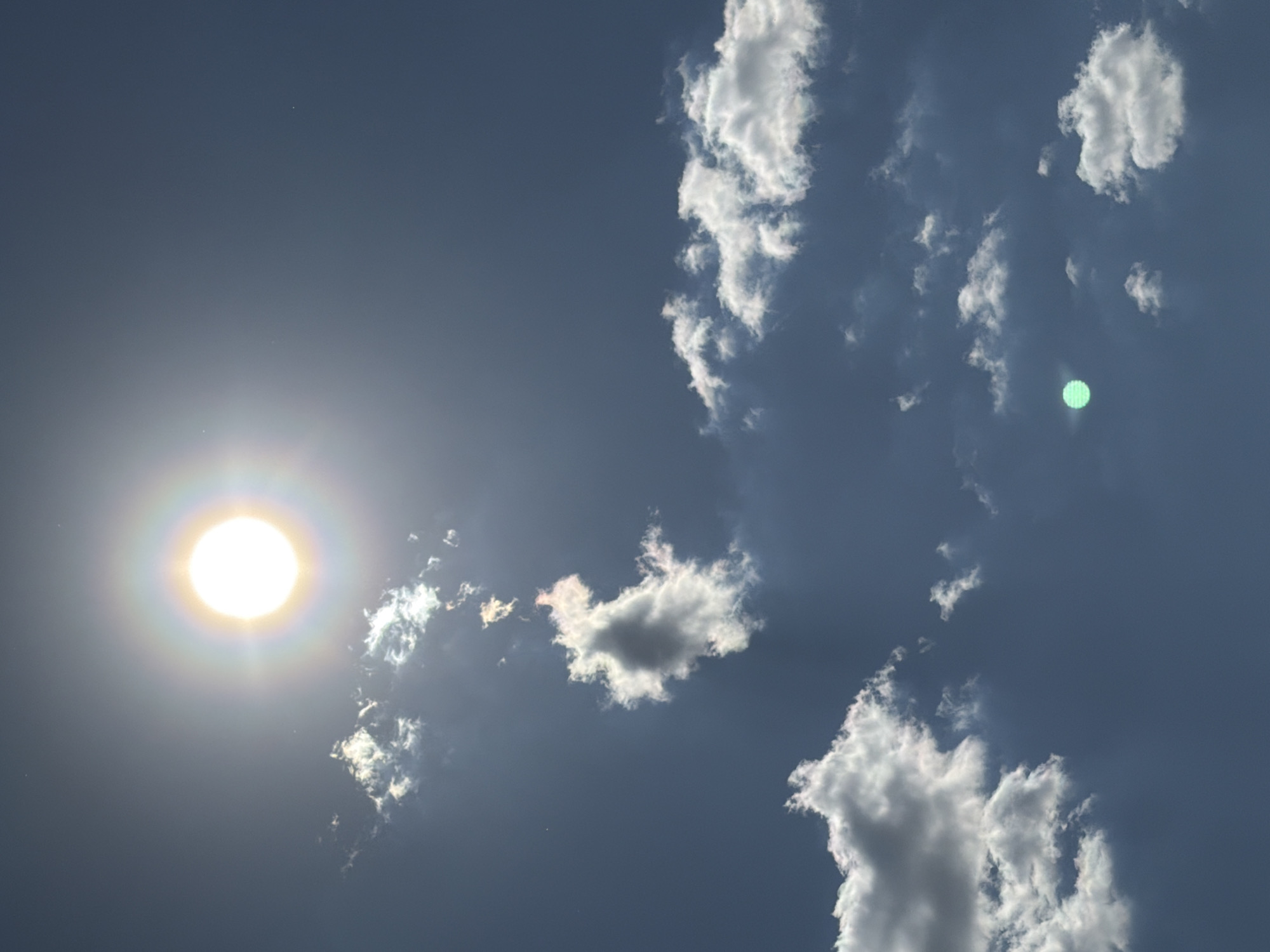 A low-angle shot of a bright sun against a blue sky with scattered cumulus clouds. The sun is on the left, and clouds are in the center and right. There is a lens flare in the clouds on the right. There is no evidence to determine that the location is in Chaikovsky, Russia. There are no people or interactions visible in the image.