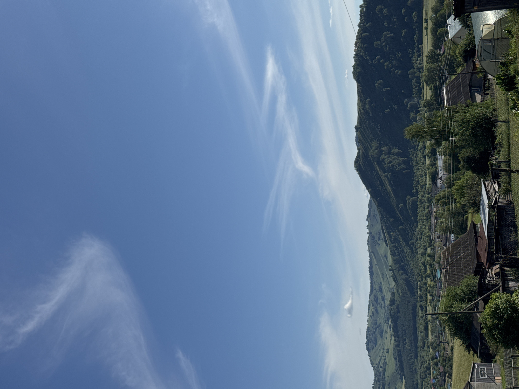 The image depicts a rural landscape on a clear day, with rolling green hills and a village nestled in the valley. The sky is a vibrant blue with wispy white clouds. In the foreground, several rustic buildings with corrugated metal roofs are visible, along with a greenhouse and a wooden fence. The village itself appears to be composed of small houses with various colored roofs, interspersed with trees and vegetation. The hills in the background are covered in a dense forest, with some lighter patches indicating meadows or clearings. The overall impression is one of a peaceful, rural setting in the town of Soloneshnoye, Russia.