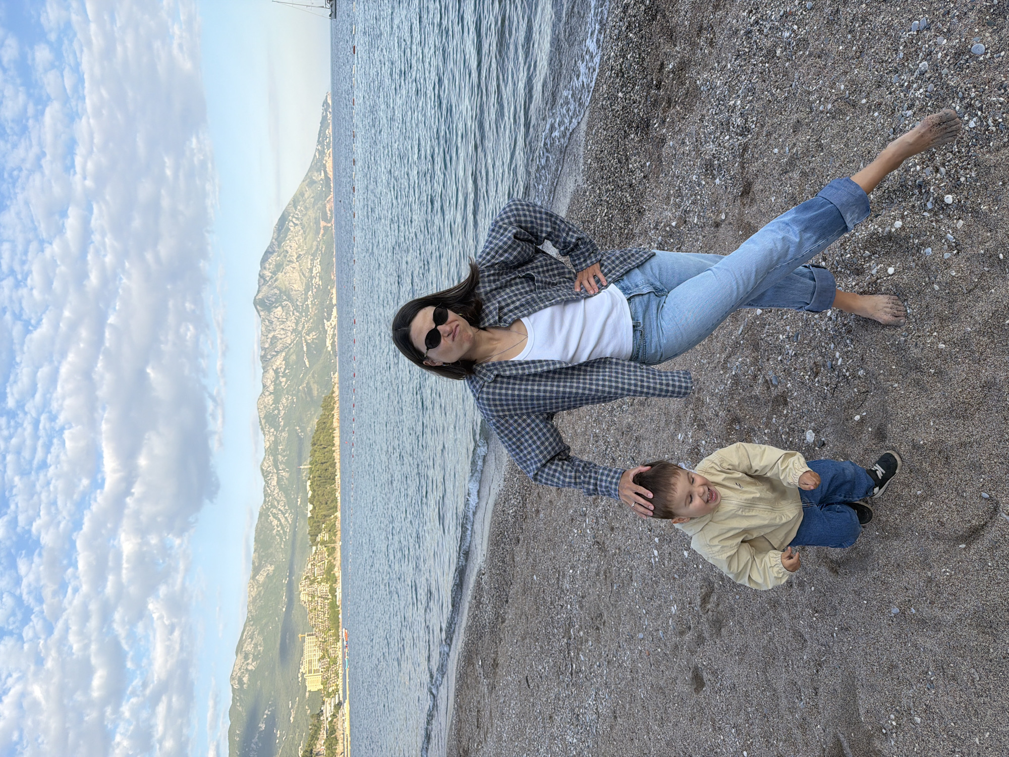 A woman and a young boy are on a pebble beach. The woman is lying on her side, facing the camera, wearing jeans, a white top, and a grey and black checkered jacket. She has sunglasses on. One leg is extended, and her bare foot is covered in sand. She is touching the boy’s head with one hand. The boy is sitting up, smiling. He is wearing a light-colored jacket and jeans, and sneakers.
The beach is located near the sea. Behind them is the coastline of Budva, Montenegro. The sky is blue with white clouds.