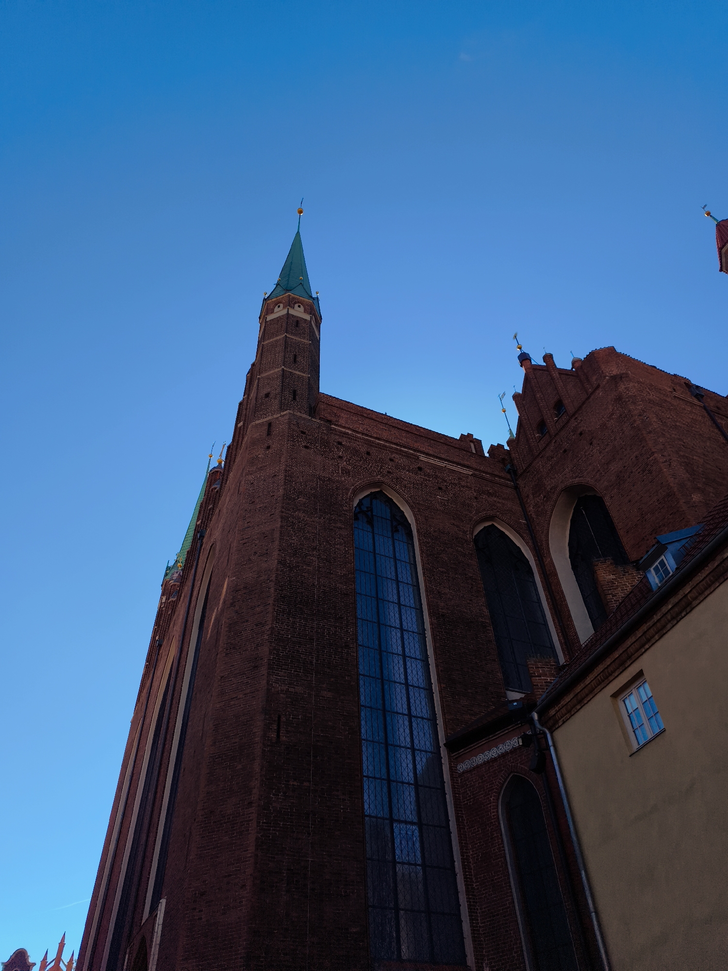 The image shows a low-angle shot of St. Mary's Church, a Gothic brick church in Gdańsk, Poland. The church is a prominent landmark with a tall, spired tower and large arched windows. The sky is a clear blue, indicating it is daytime. The facade of the church is made of dark red brick. A section of a yellow building is visible to the right of the church, with a small window. The image is taken from a street level perspective, looking up towards the sky.